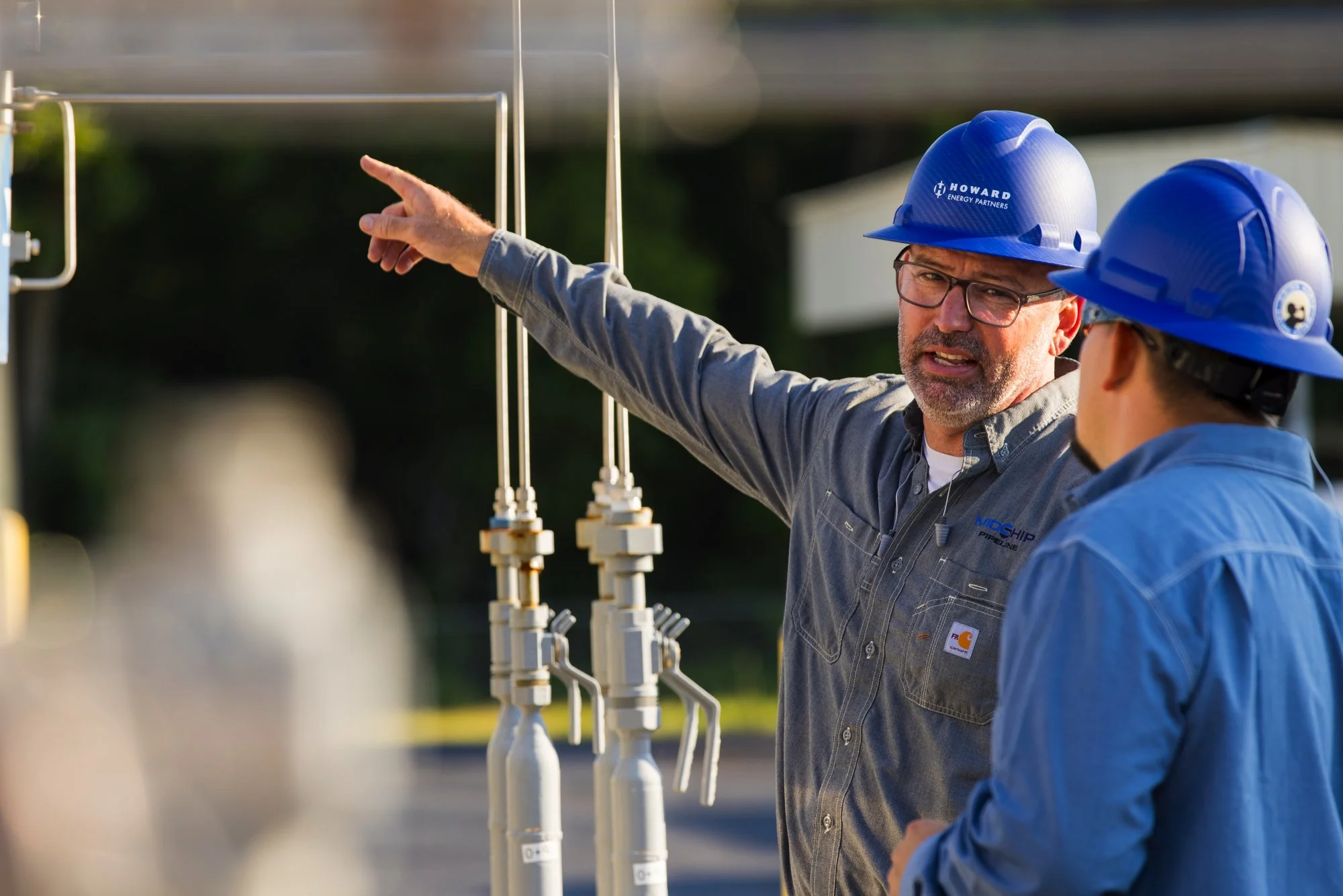 Two male workers wearing blue safety helmets and work shirts in conversation outdoors near industrial pipes and valves, with one pointing.