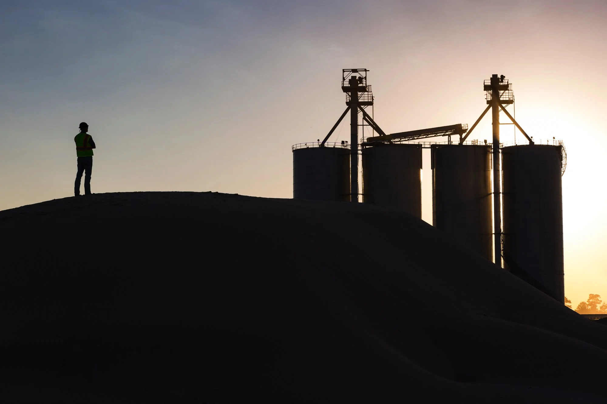 A silhouette of a person in a safety vest standing on a mound looking at industrial silos during sunset.