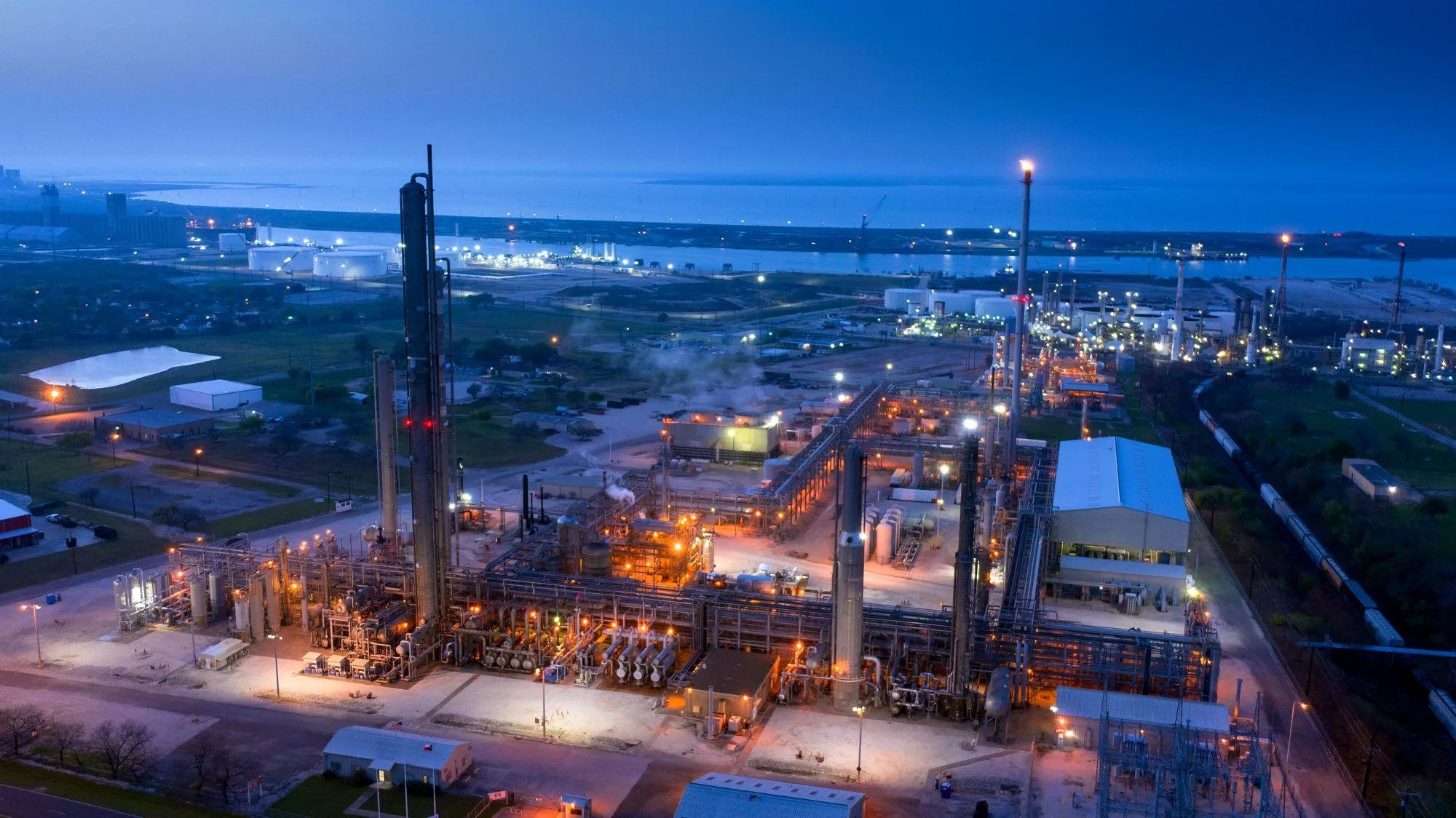 An industrial refinery at dusk with illuminated equipment, pipelines, and storage tanks, near a body of water.