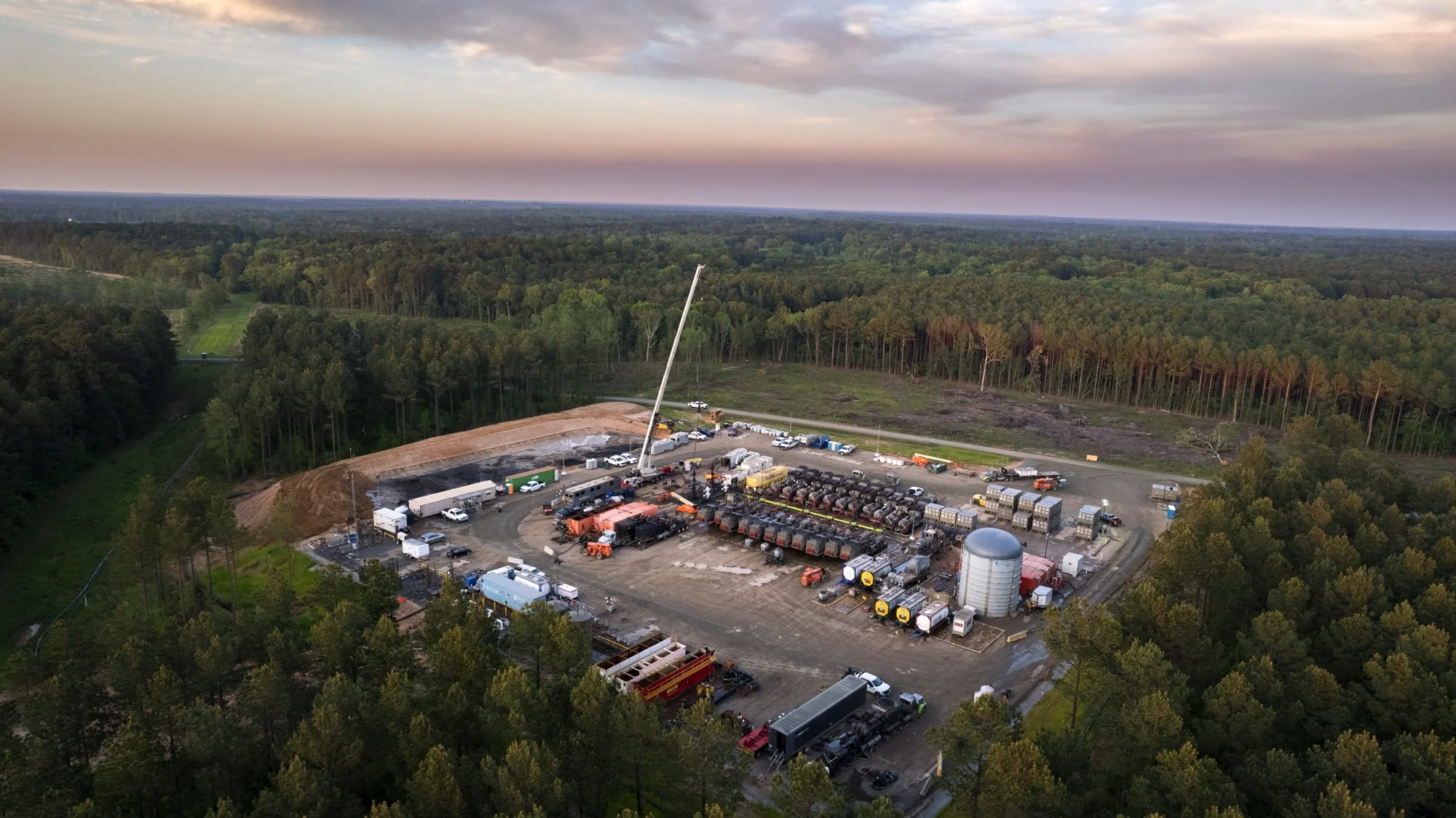 Aerial view of an industrial site or construction area surrounded by dense forest, with equipment, vehicles, and structures including a large silos, containers, and fuel tanks, and a crane.