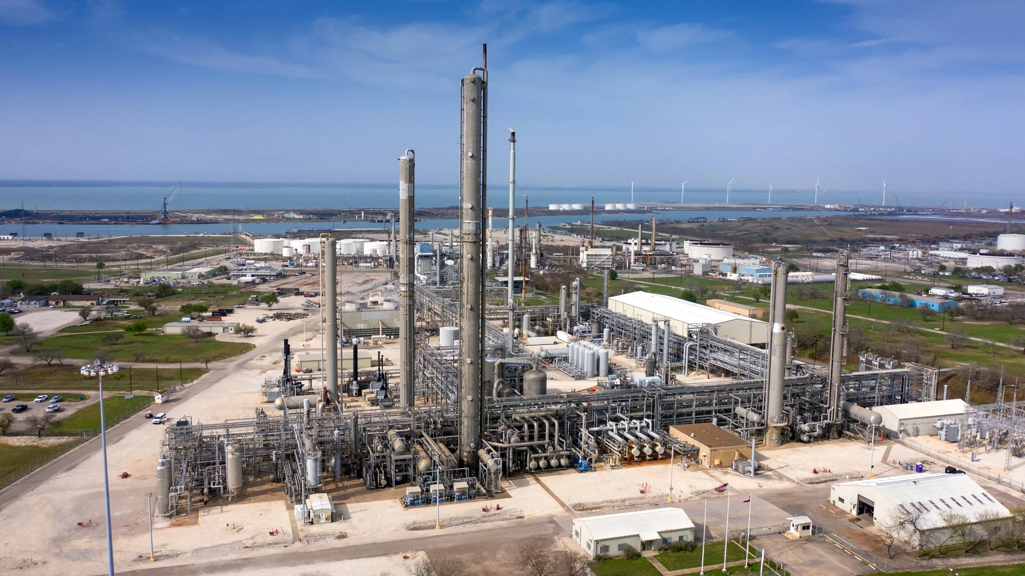 Industrial plant with multiple pipes and tall smokestacks near a body of water, with wind turbines in the distance.