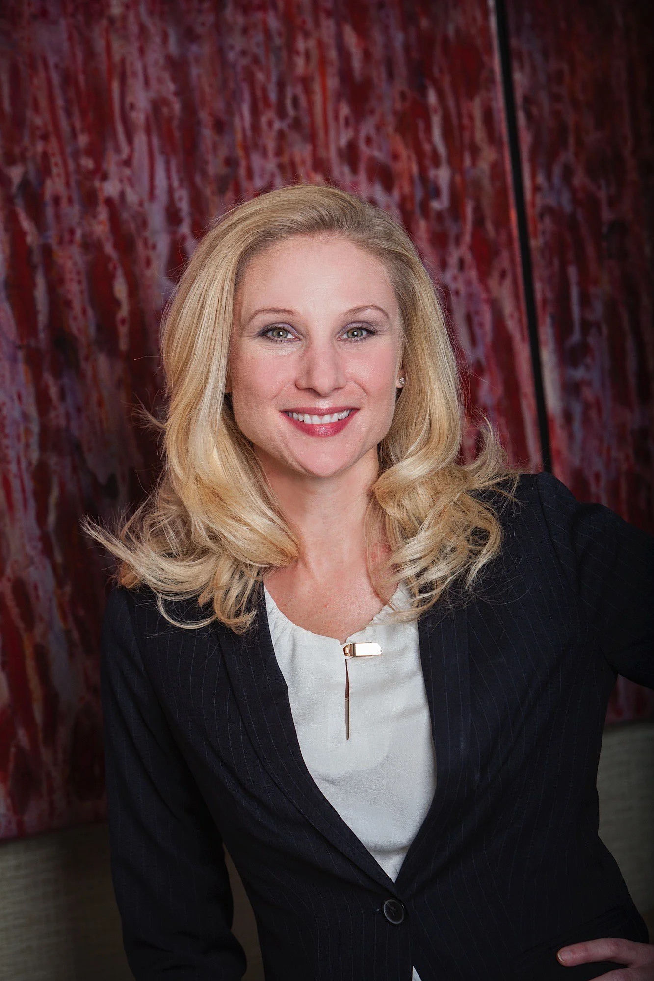 A smiling woman with blonde hair in business attire, posing in front of an abstract red and black background.