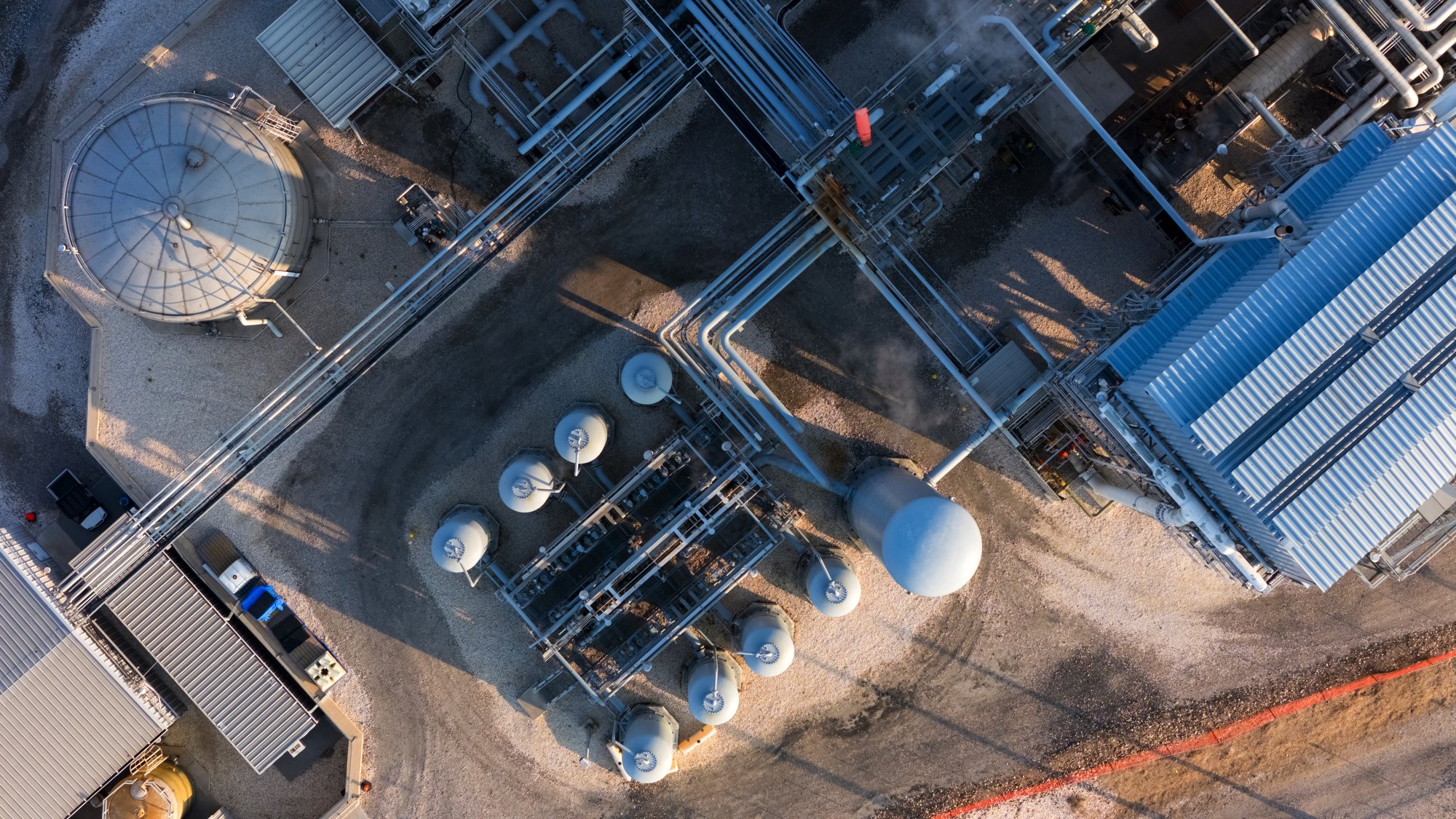 An aerial view of an industrial facility with large metal storage tanks, pipelines, and structures, with shadows cast on the gravel ground.