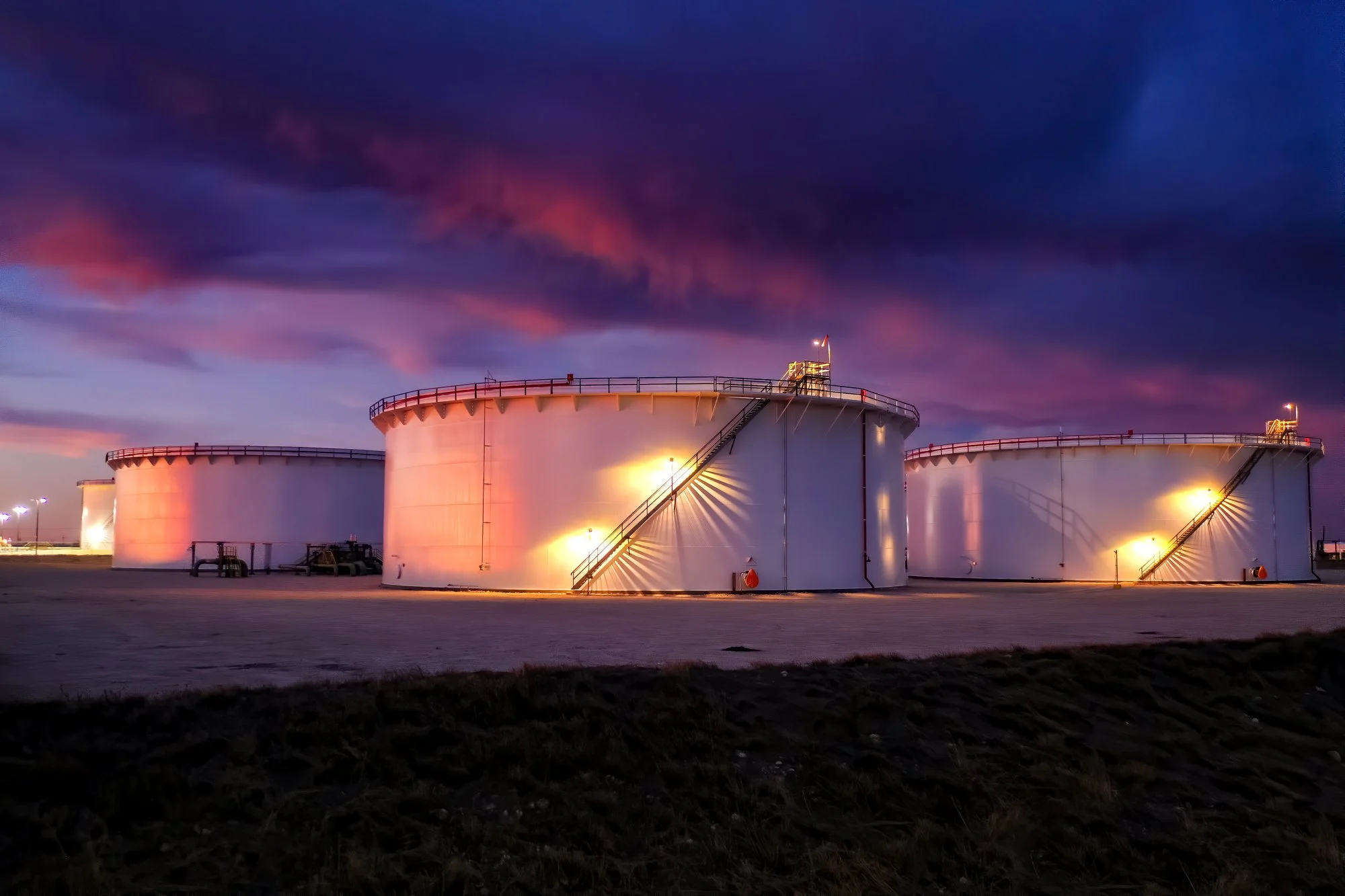 Three large white industrial storage tanks at dusk with colorful pink and purple clouds in the sky and ground in the foreground.