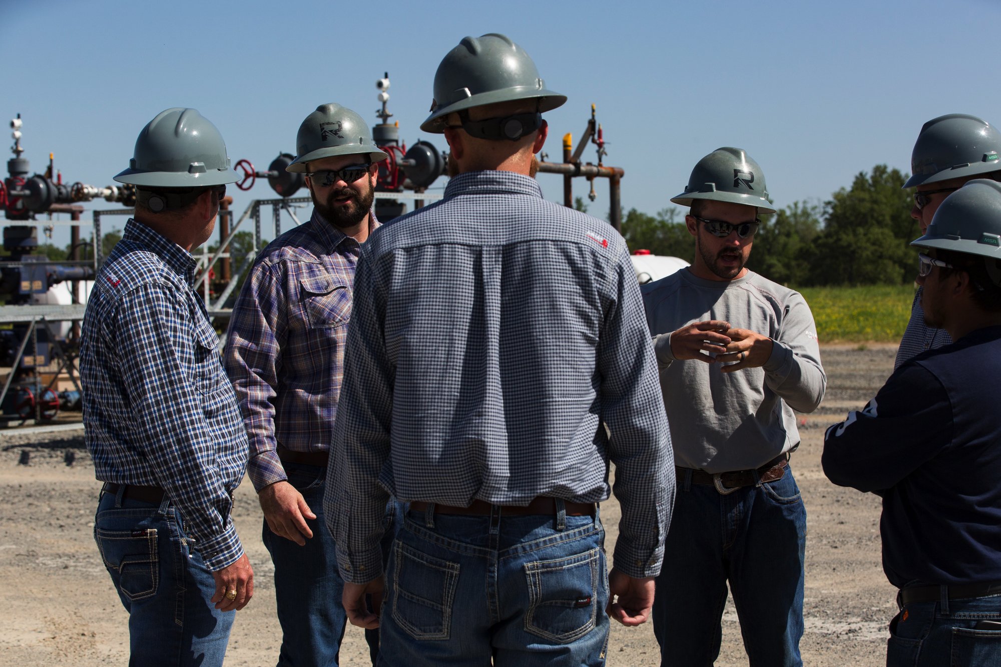 Group of six men wearing hard hats and casual clothing standing outdoors near industrial equipment, engaged in a conversation.