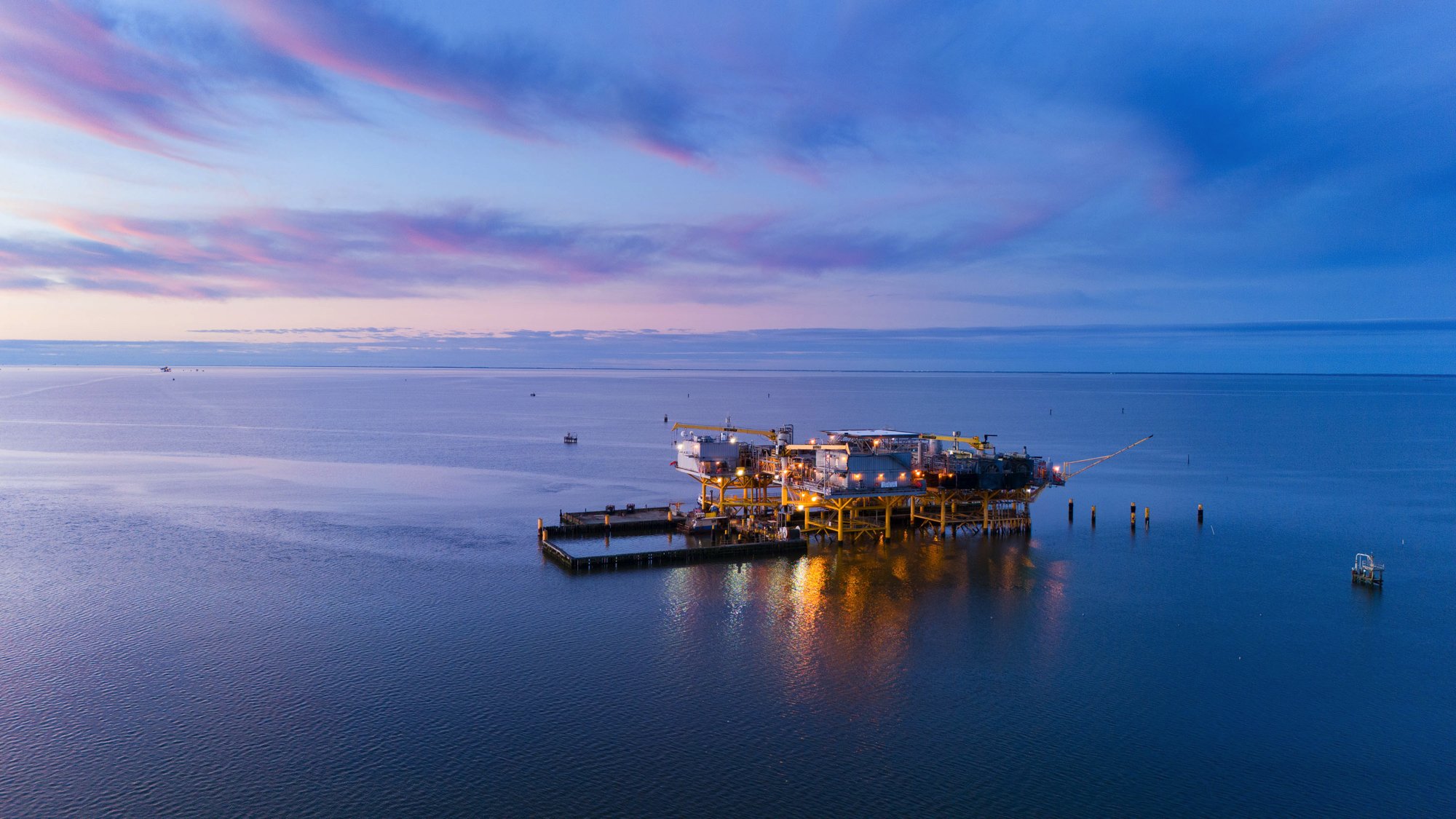 An offshore oil platform illuminated by lights floats in a calm sea at dusk, with a colorful sky overhead.