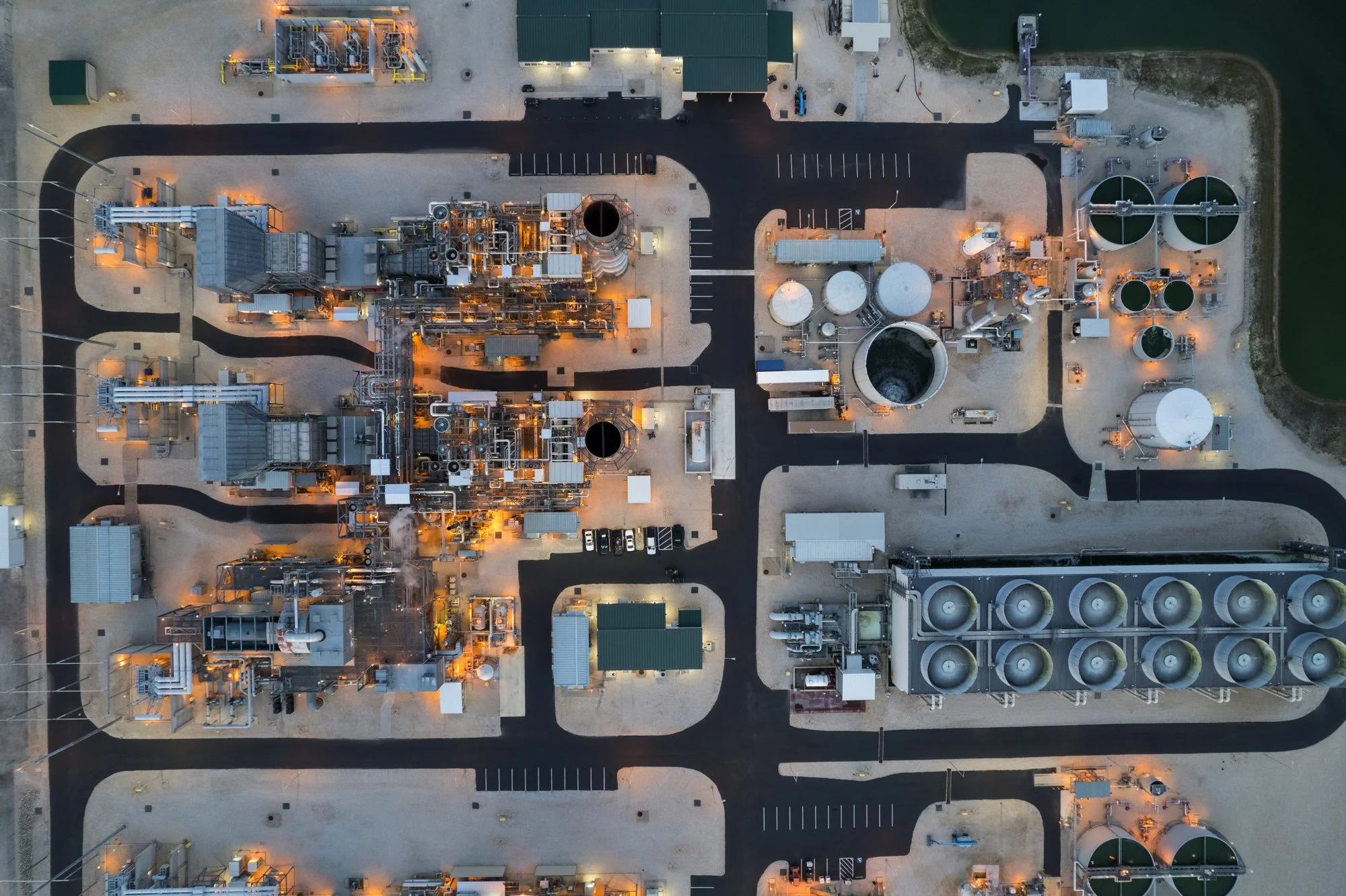 An overhead aerial view of an industrial facility with tanks, equipment, and pipelines, surrounded by paved roads and parking areas.
