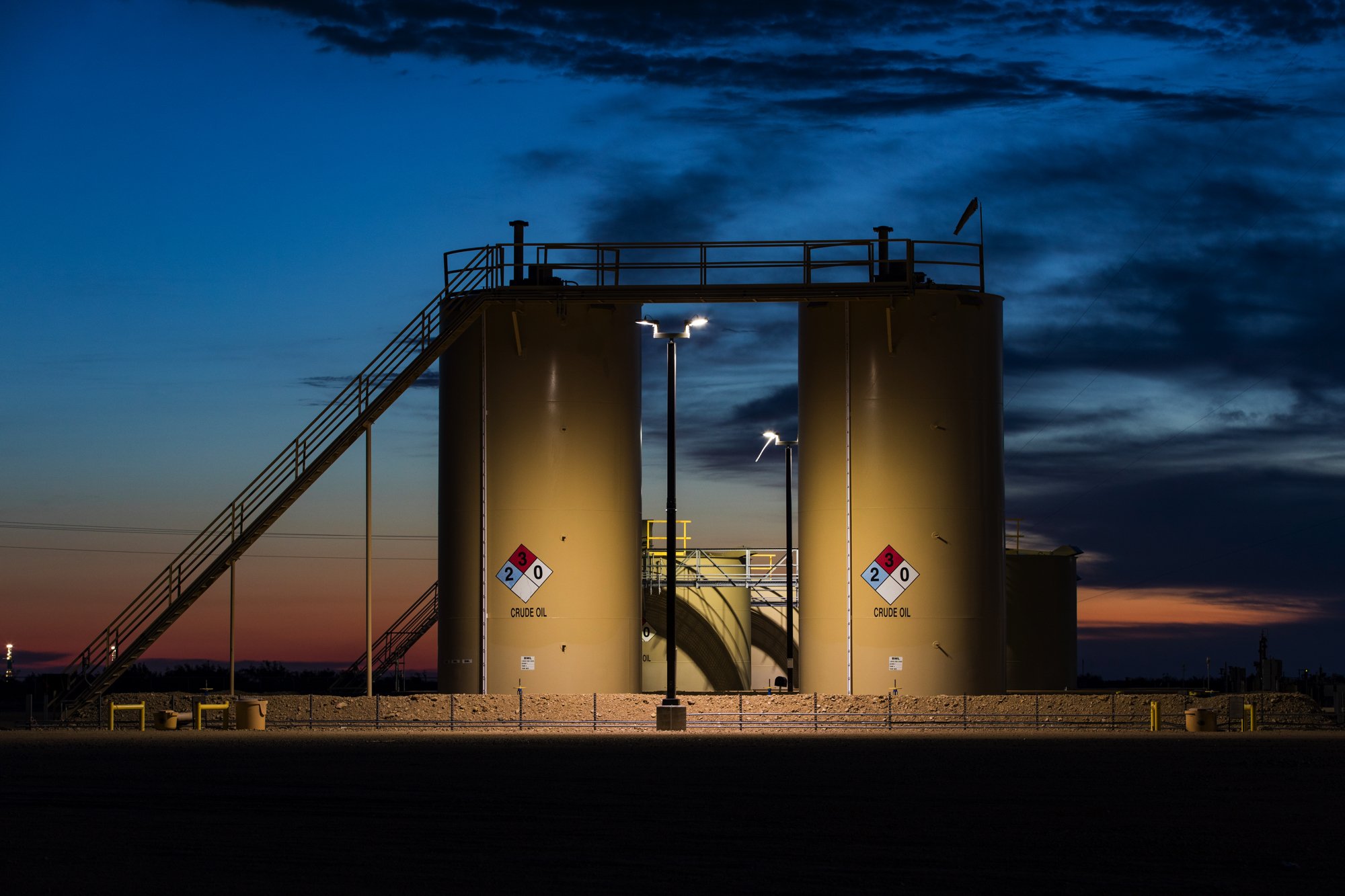 Industrial storage tanks labeled 'Crude Oil' with hazard signs, illuminated at dusk, with a ladder leading to the top and a sky with clouds in the background.