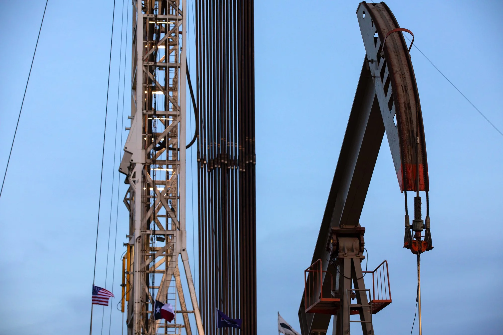 Close-up view of construction crane and oil pumpjack against a blue sky with flags at the bottom.