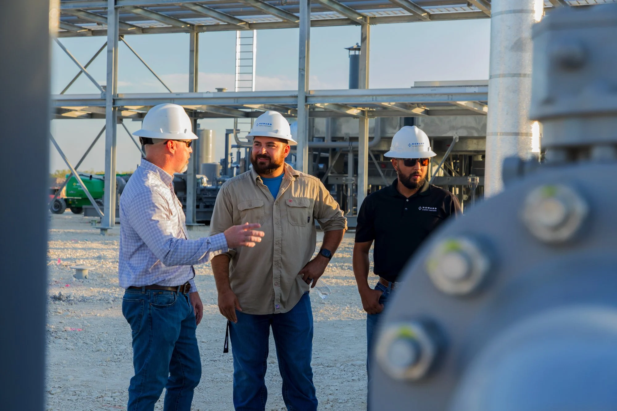 Three construction workers wearing helmets and safety glasses talking at an industrial site with infrastructure and equipment in the background.