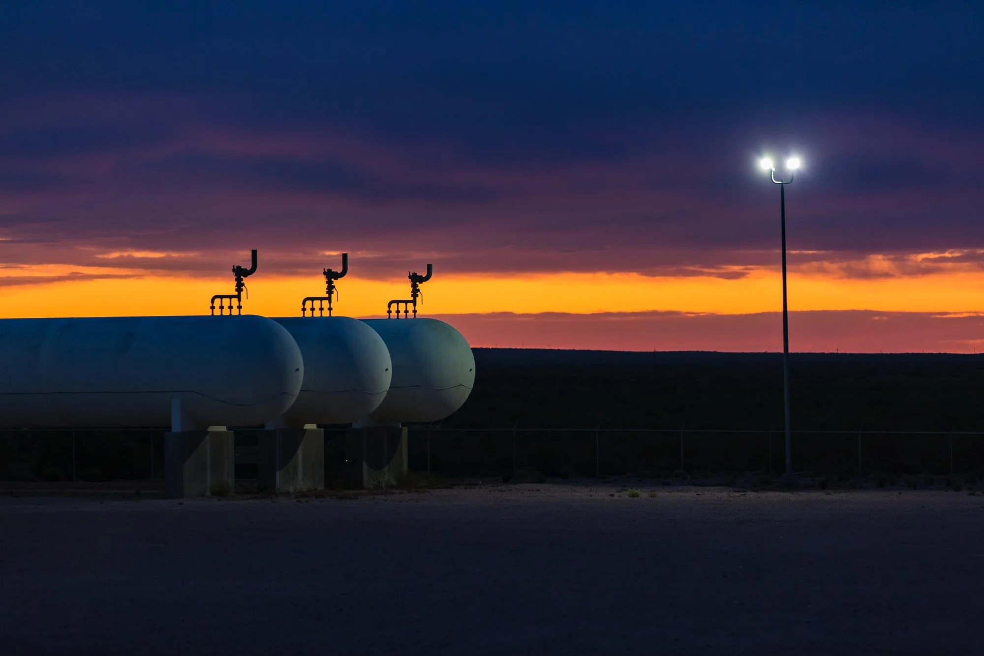 Three large white storage tanks with black pipes on top, illuminated by a tall outdoor light at dusk, with a colorful sunset sky in the background.