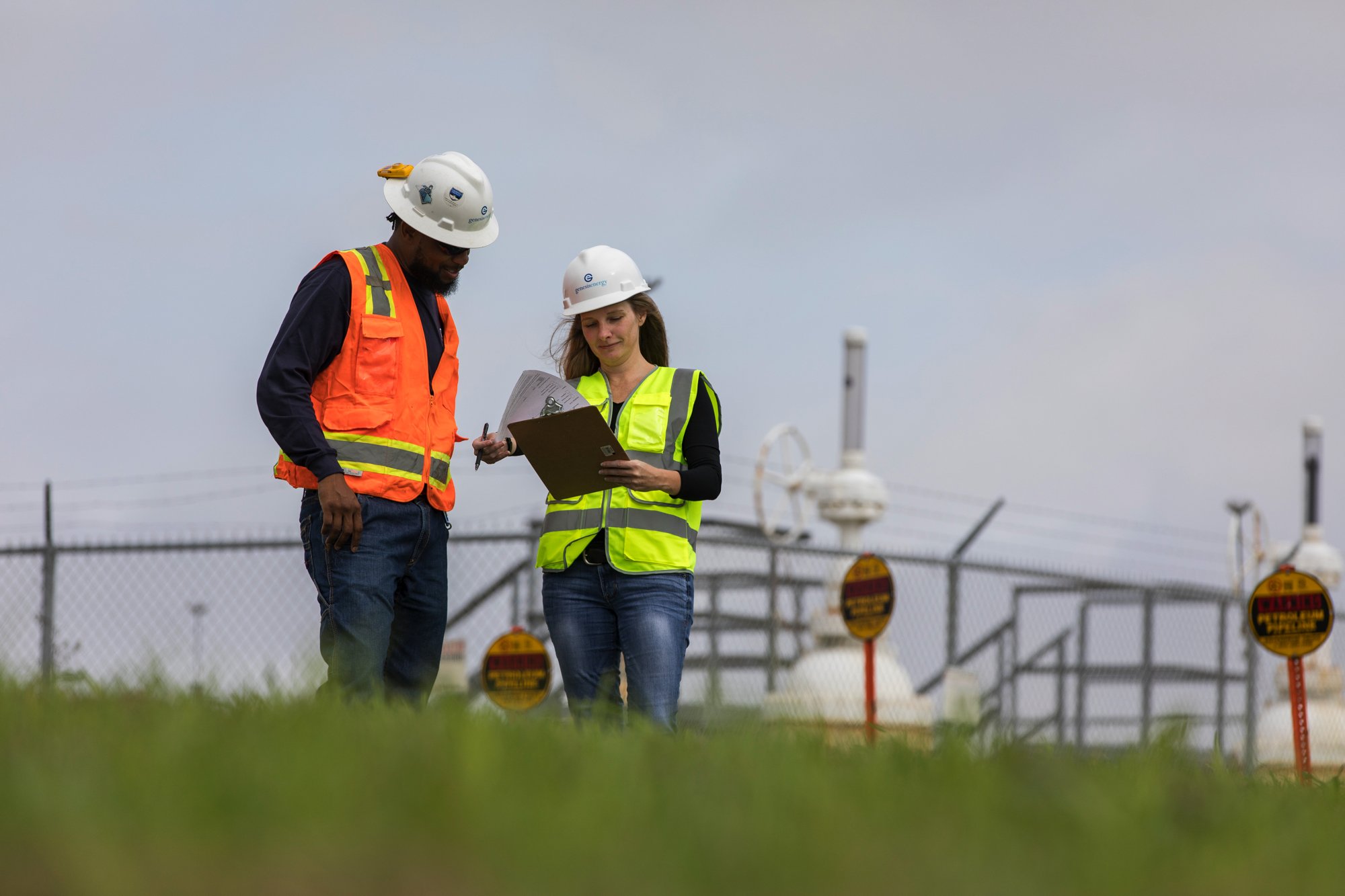 Two workers in safety vests and helmets standing outdoors near industrial equipment, reviewing documents.