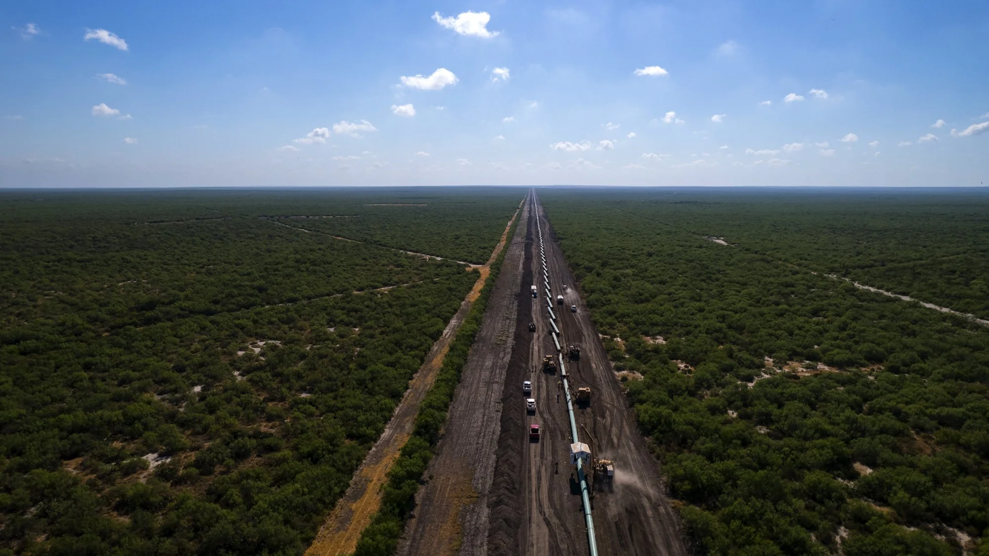An aerial view of a long pipeline construction through a green landscape with scattered bushes under a partly cloudy sky.