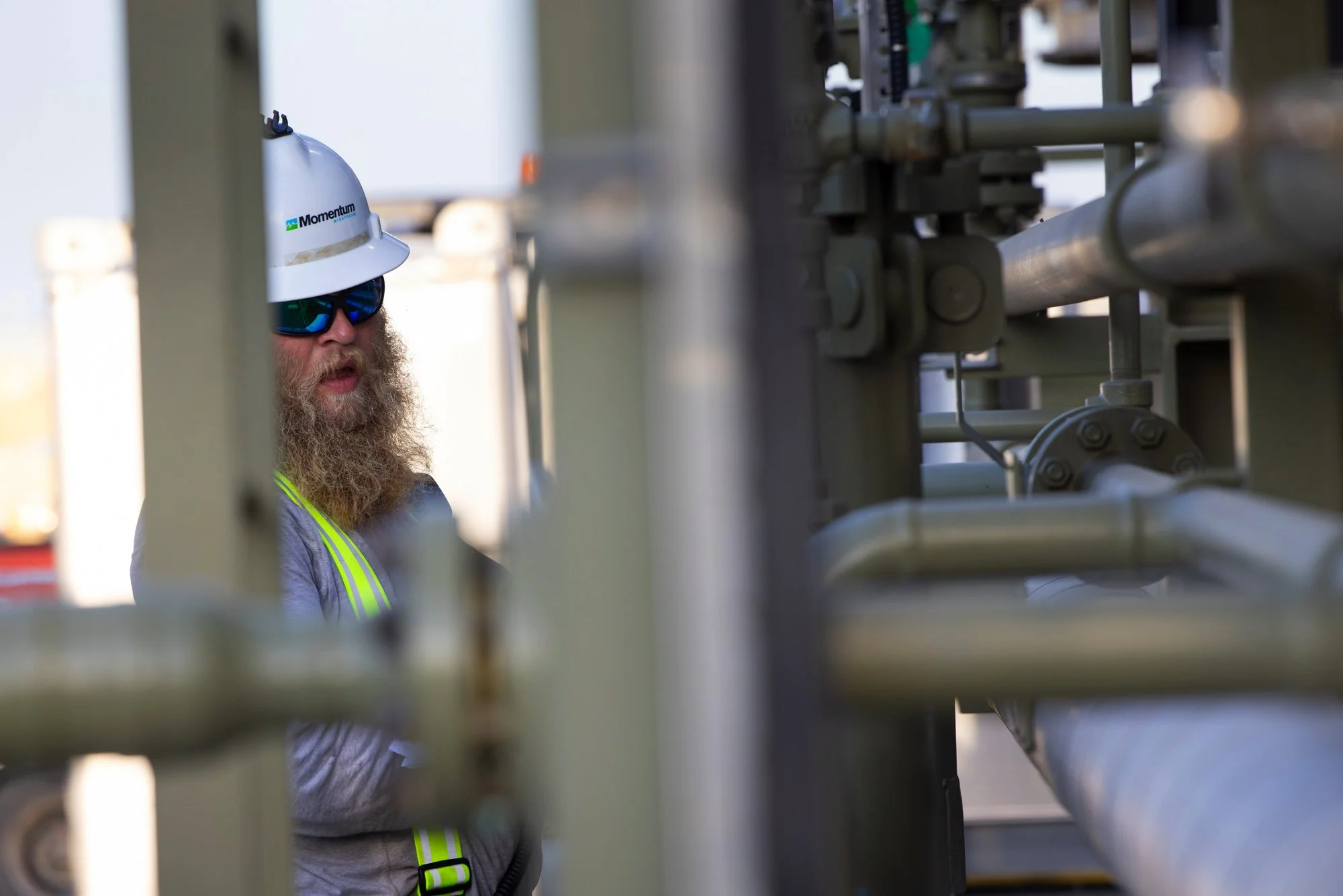 A bearded man wearing a white safety helmet with a logo, sunglasses, and a gray safety vest with yellow stripes stands behind metal industrial pipes and equipment.