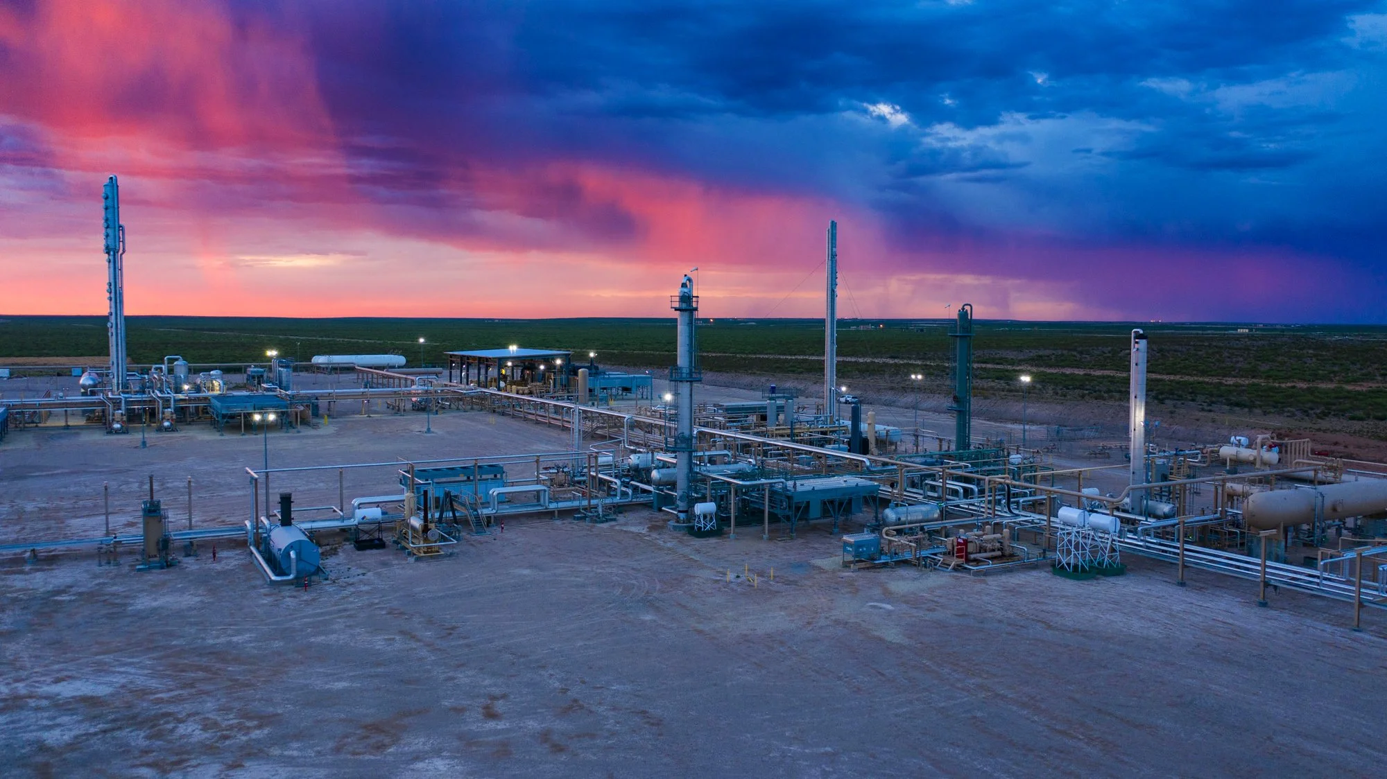 An oil or natural gas processing plant at sunset with a colorful sky in the background. The facility has multiple pipelines, tanks, and industrial structures.