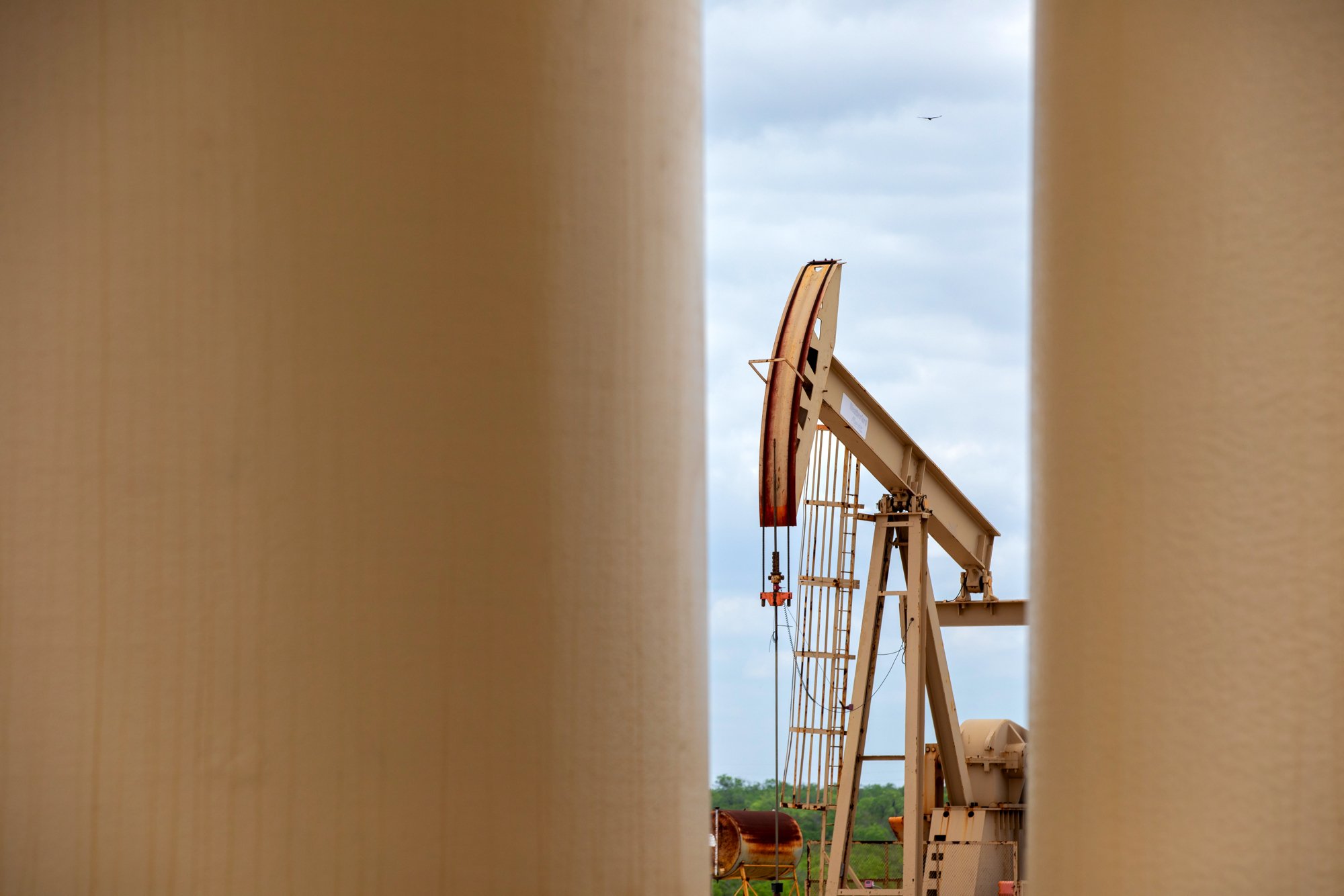 An oil pumpjack seen through a narrow gap between two vertical beige-colored objects. The pumpjack is brown and white, with a curved counterweight and a walking beam, set against a partly cloudy sky.
