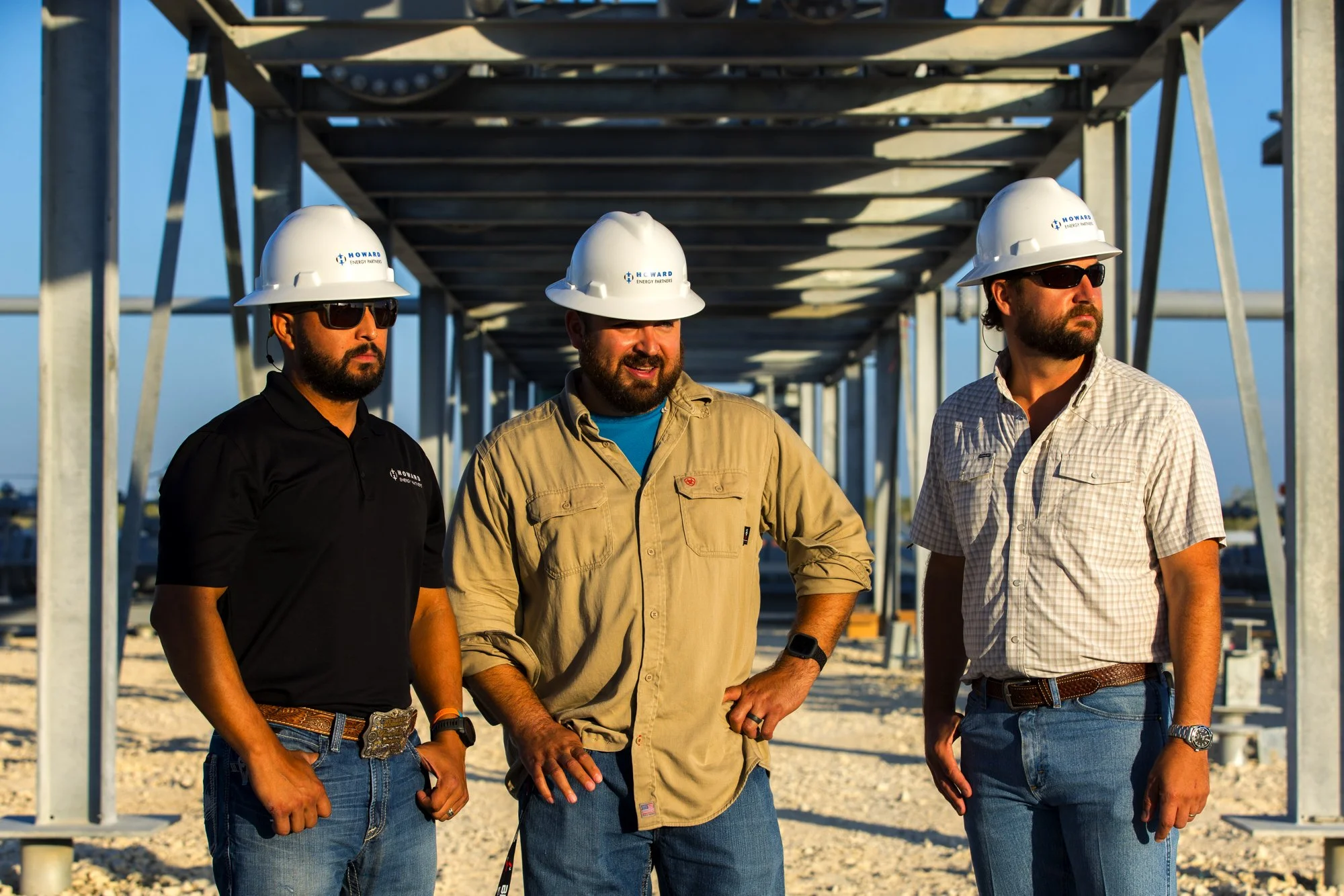 Three construction workers standing under metal scaffolding at a construction site, wearing hard hats and safety glasses.