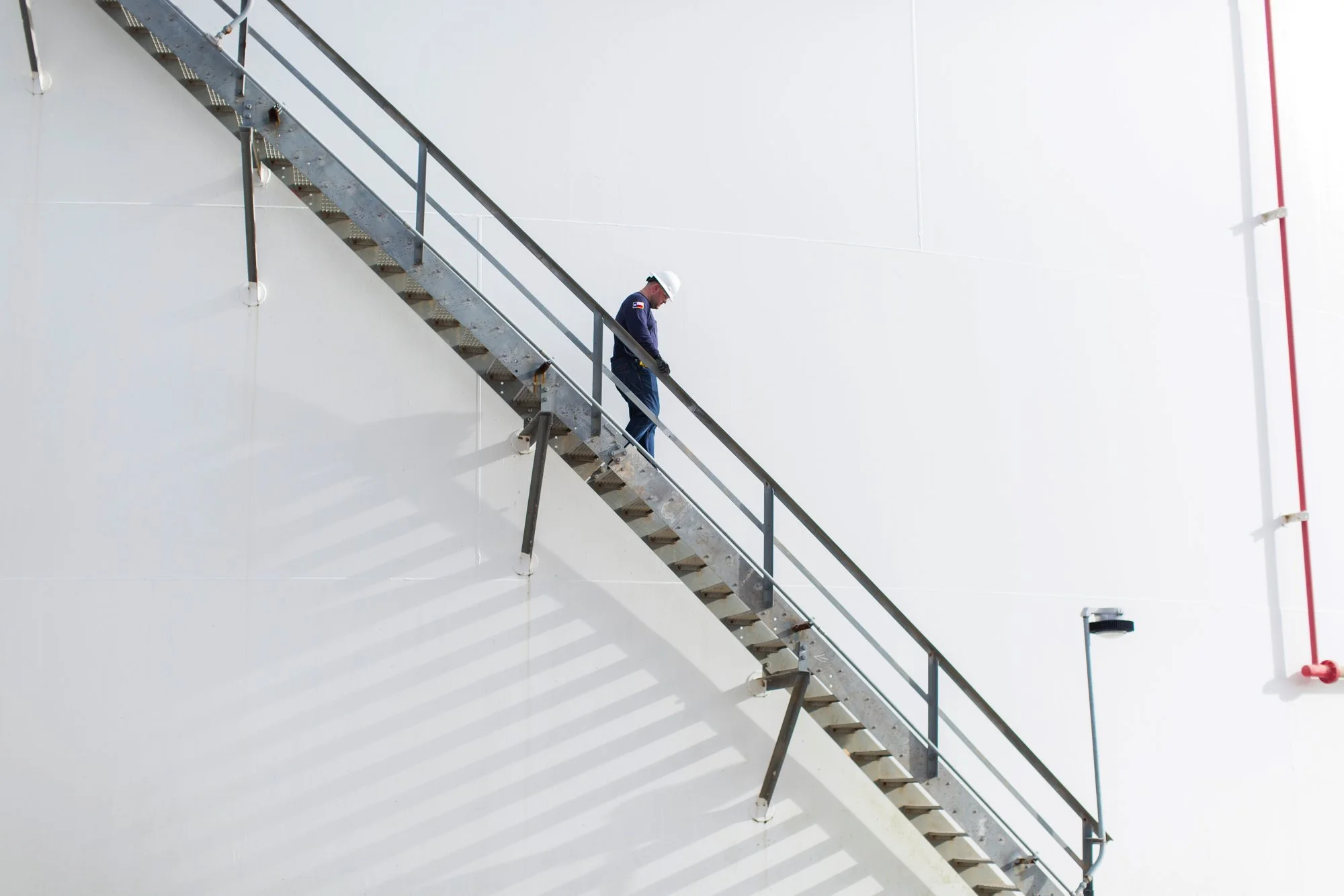 A worker in a safety helmet and uniform walking down an outdoor metal staircase attached to a white building wall.