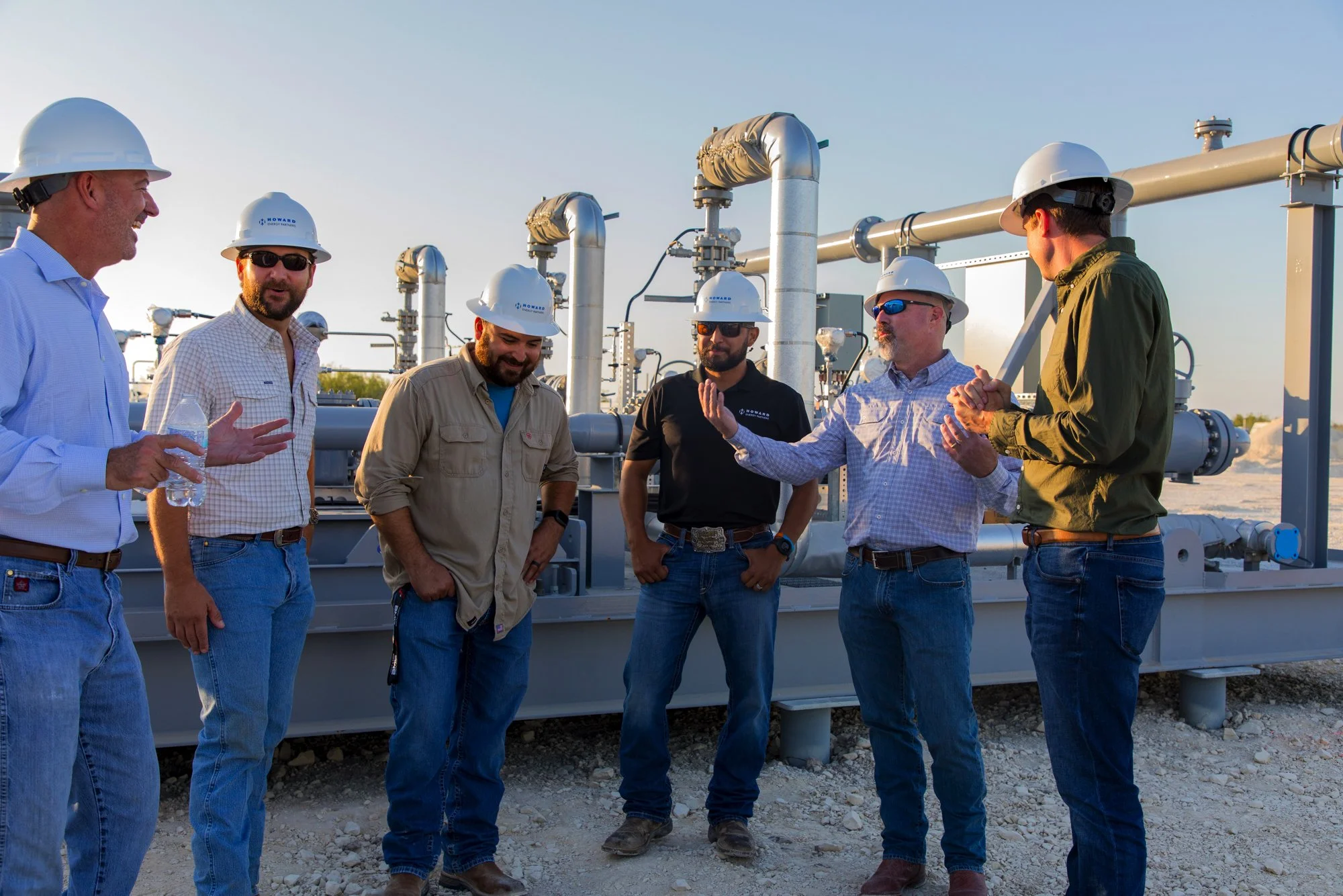Six men in hard hats and casual clothing standing in front of industrial pipes and equipment outdoors, having a discussion.