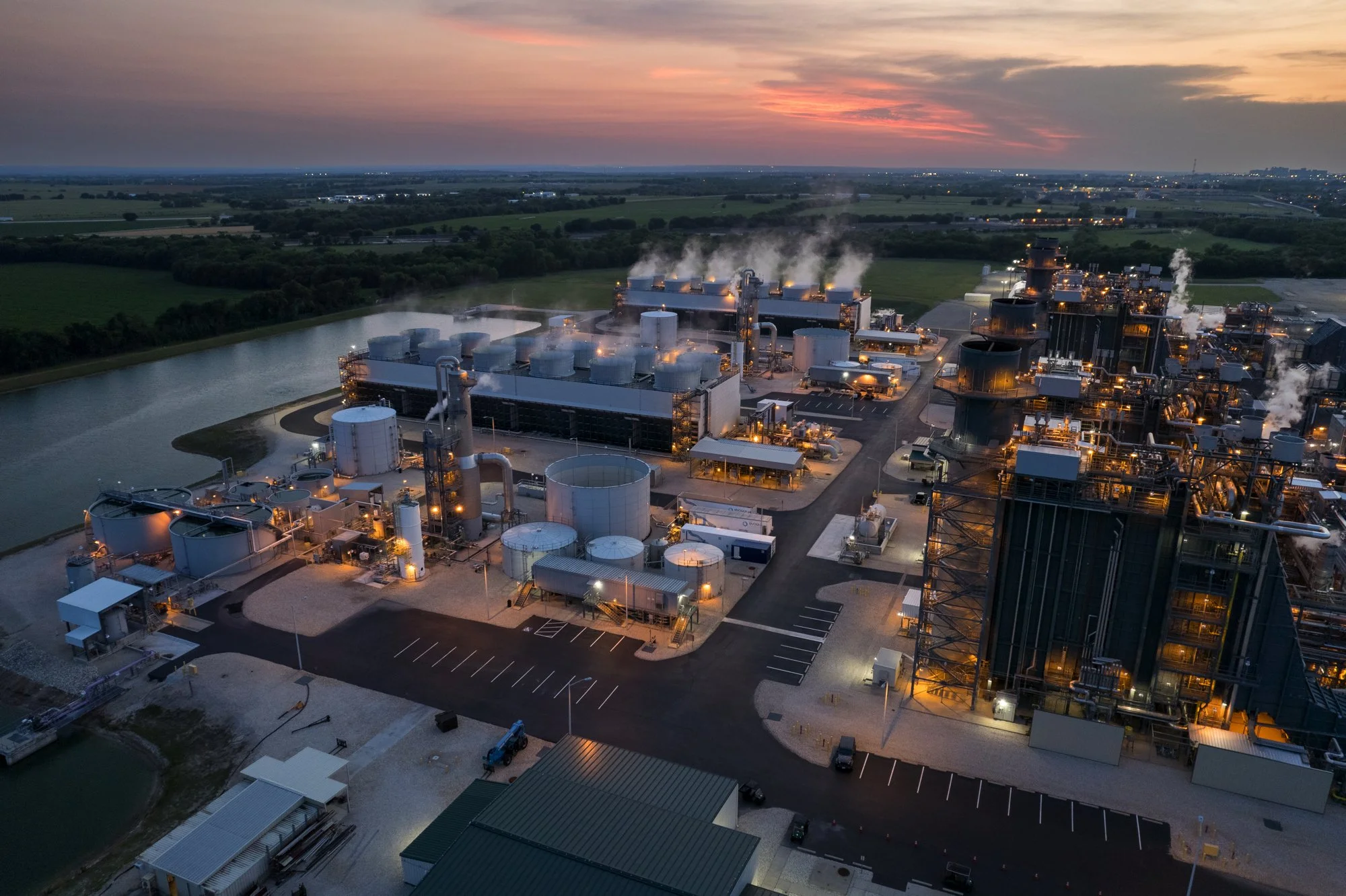 An aerial view of an industrial factory complex during sunset, with numerous buildings, tanks, pipes, and steam emissions, surrounded by open fields and a water body.