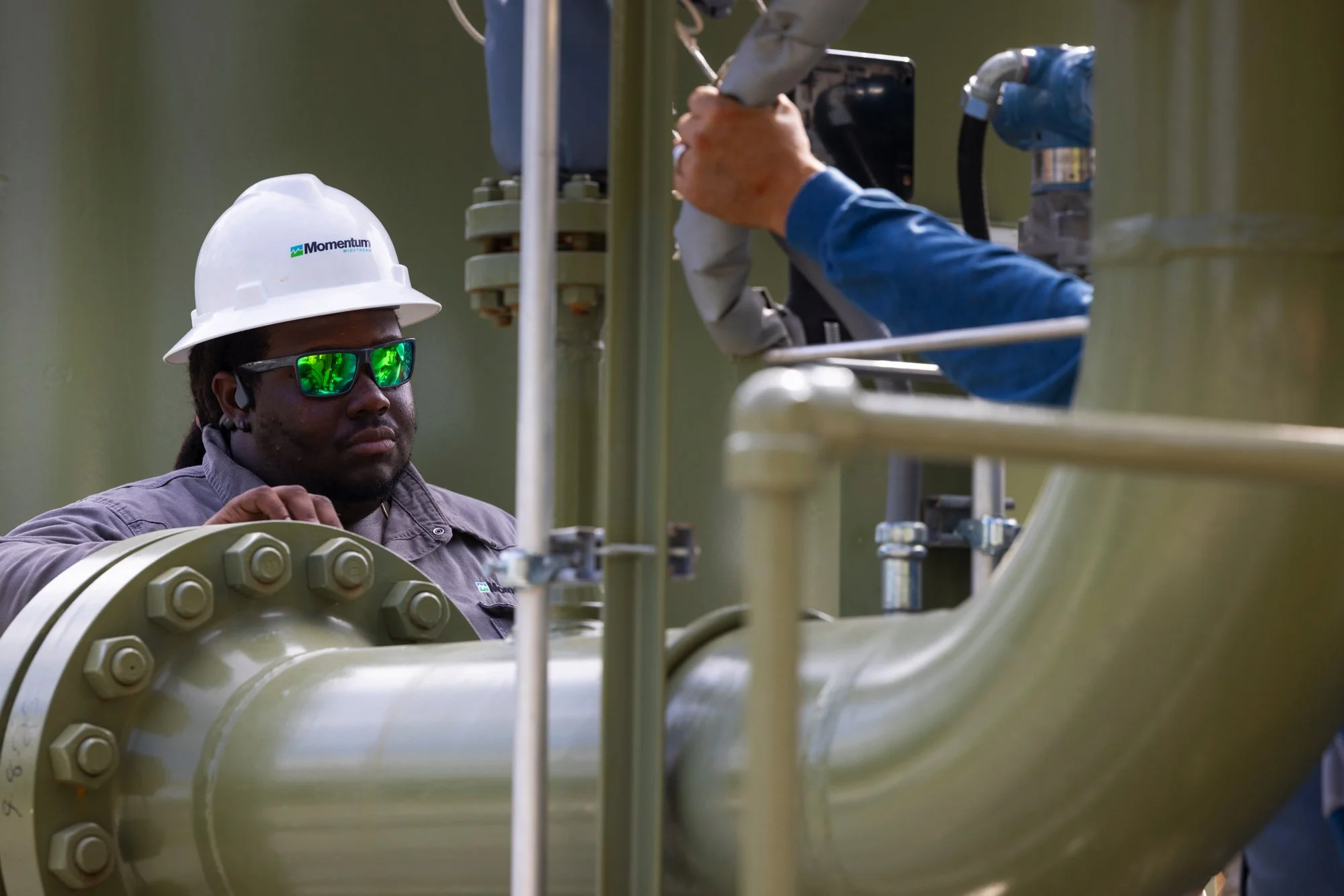 Two workers inspecting a large industrial pipe; one wearing a white hard hat with a logo and green reflective sunglasses, the other wearing a blue shirt and gloves, adjusting equipment.