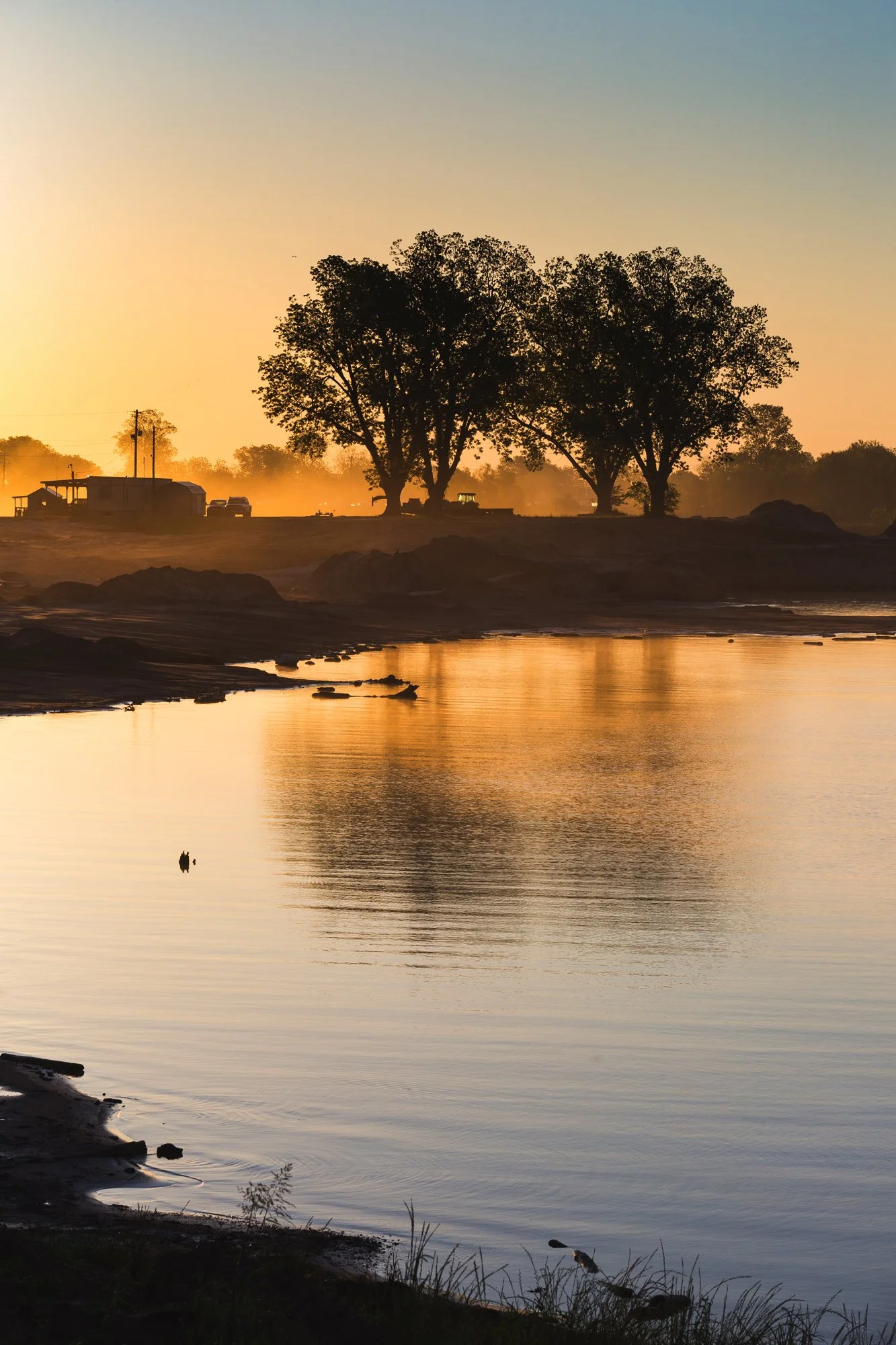 Sunset or sunrise over a calm river with trees in silhouette on the far bank and a small structure and vehicles in the background.