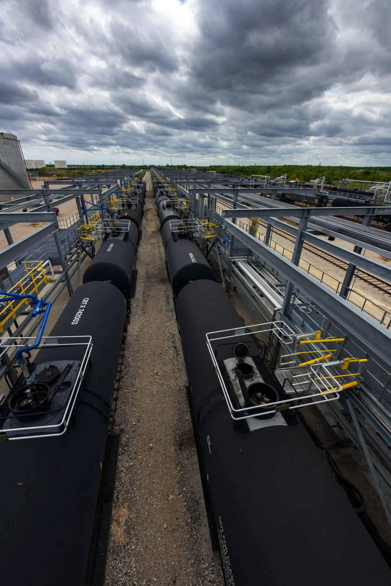 Aerial view of multiple black tanker trains on parallel tracks under a cloudy sky, with industrial structures and green landscape in the background.
