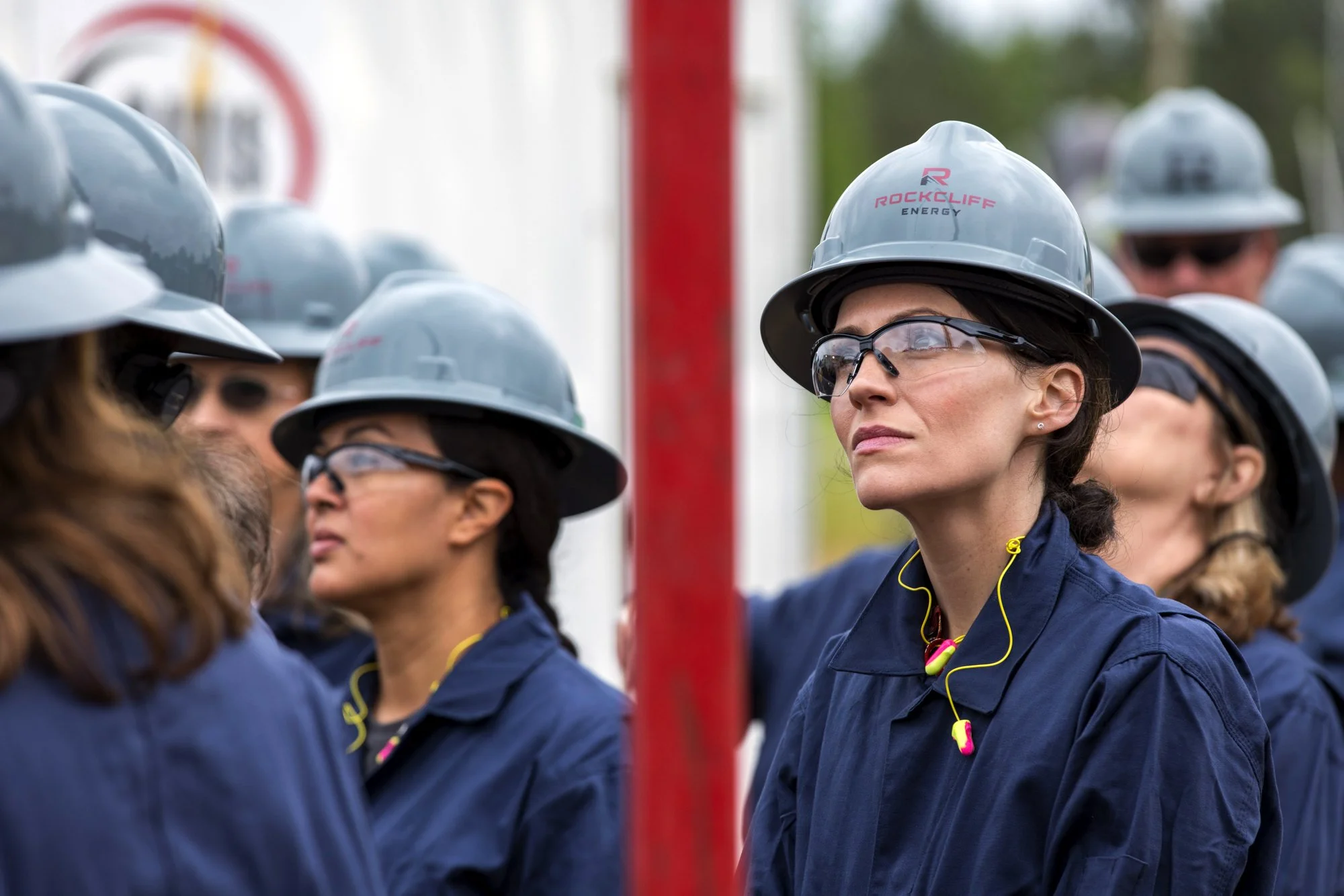 Group of women in blue uniforms and gray safety helmets at an outdoor work site, with one woman in focus wearing glasses and a yellow headset, listening attentively.