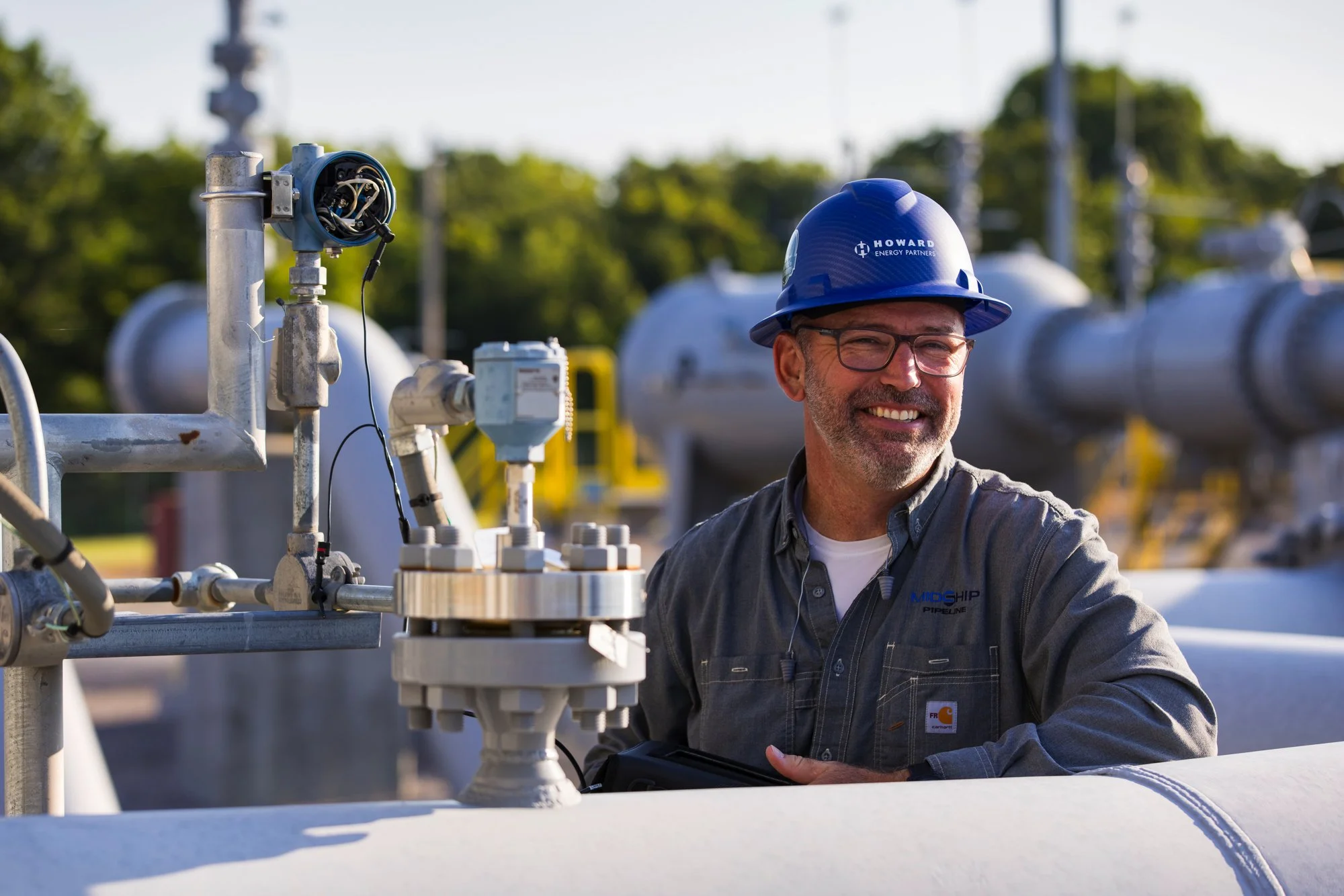 A smiling man wearing glasses, a blue hard hat, and a gray work shirt standing near industrial pipeline equipment outdoors.