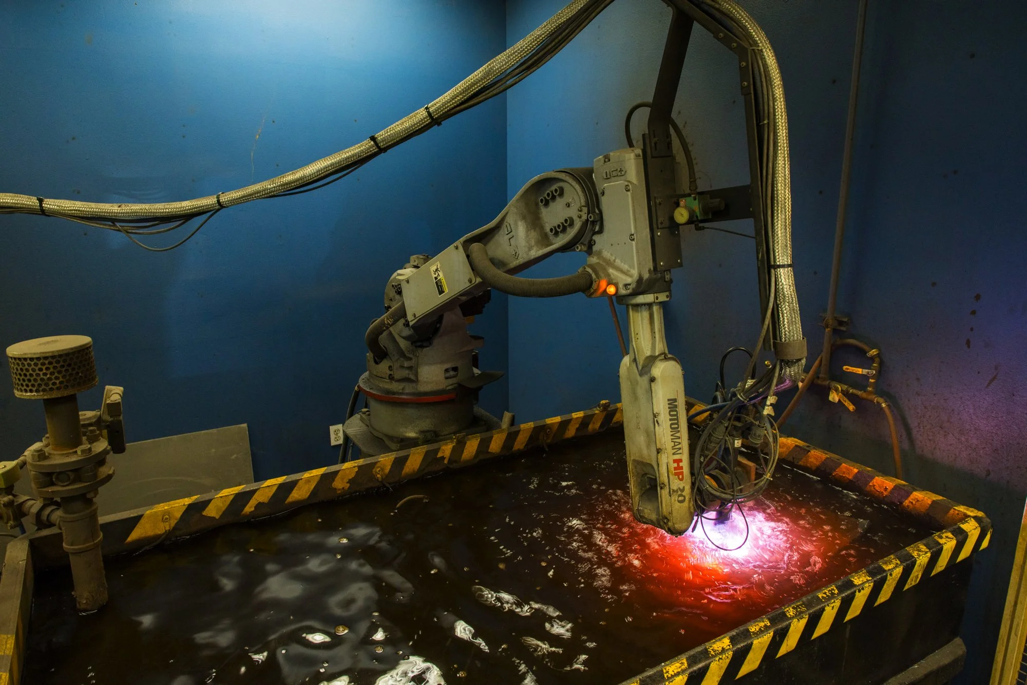 Industrial robotic arm welding or cutting in a blue-walled workshop, with a black container holding liquid beneath it.