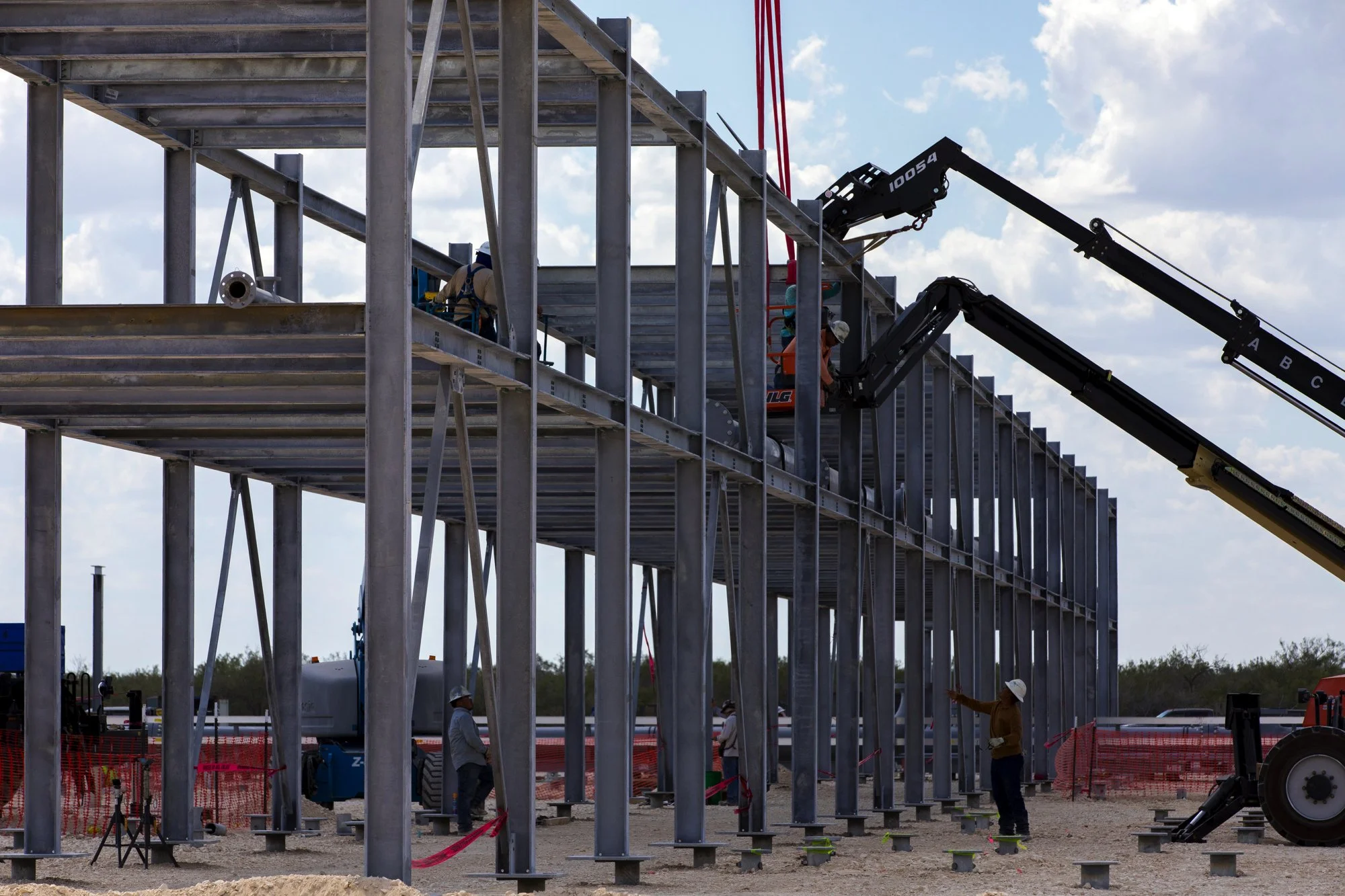 Construction workers assemble a steel building frame using cranes on a construction site under a partly cloudy sky.