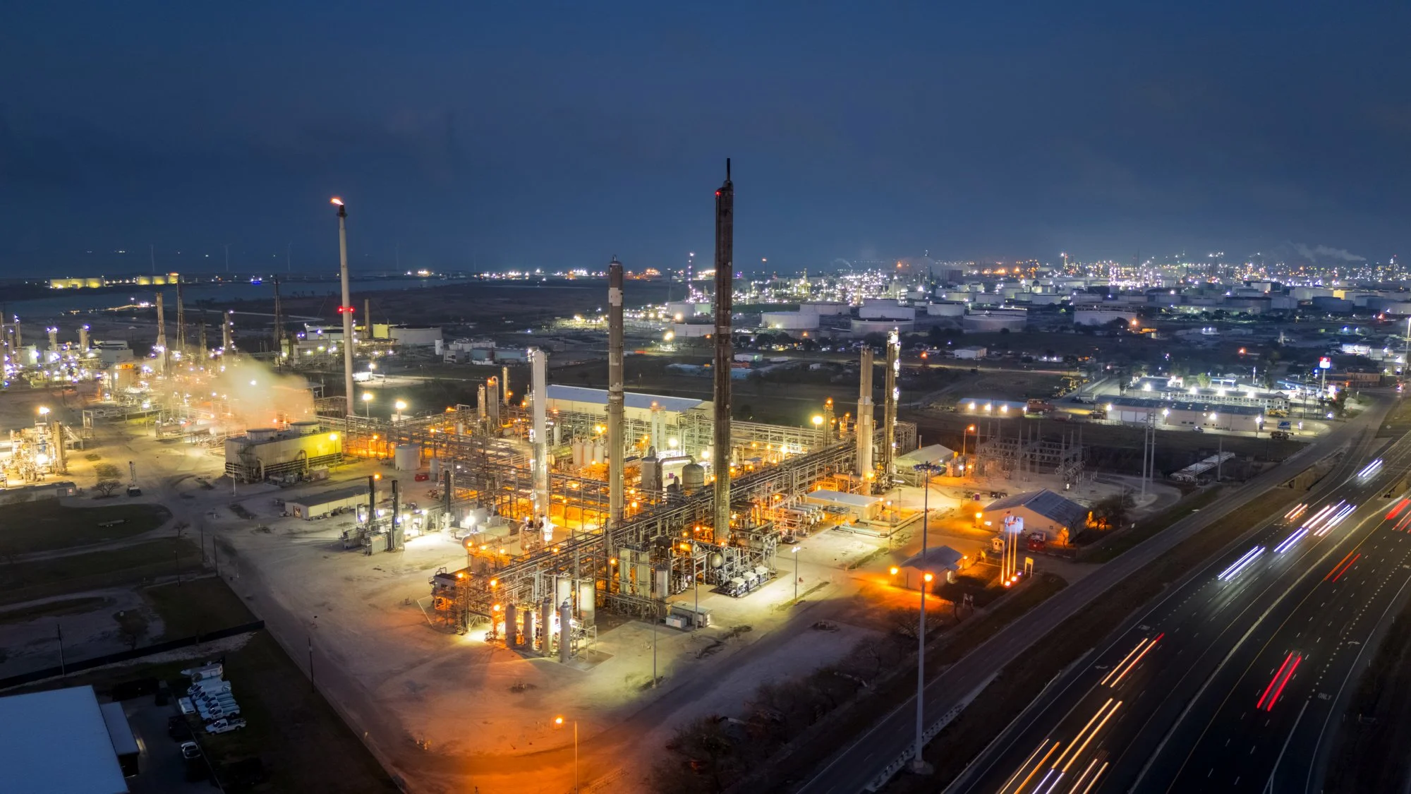 Night view of an industrial oil refinery with tall columns and bright lights, adjacent to a highway with moving vehicles.