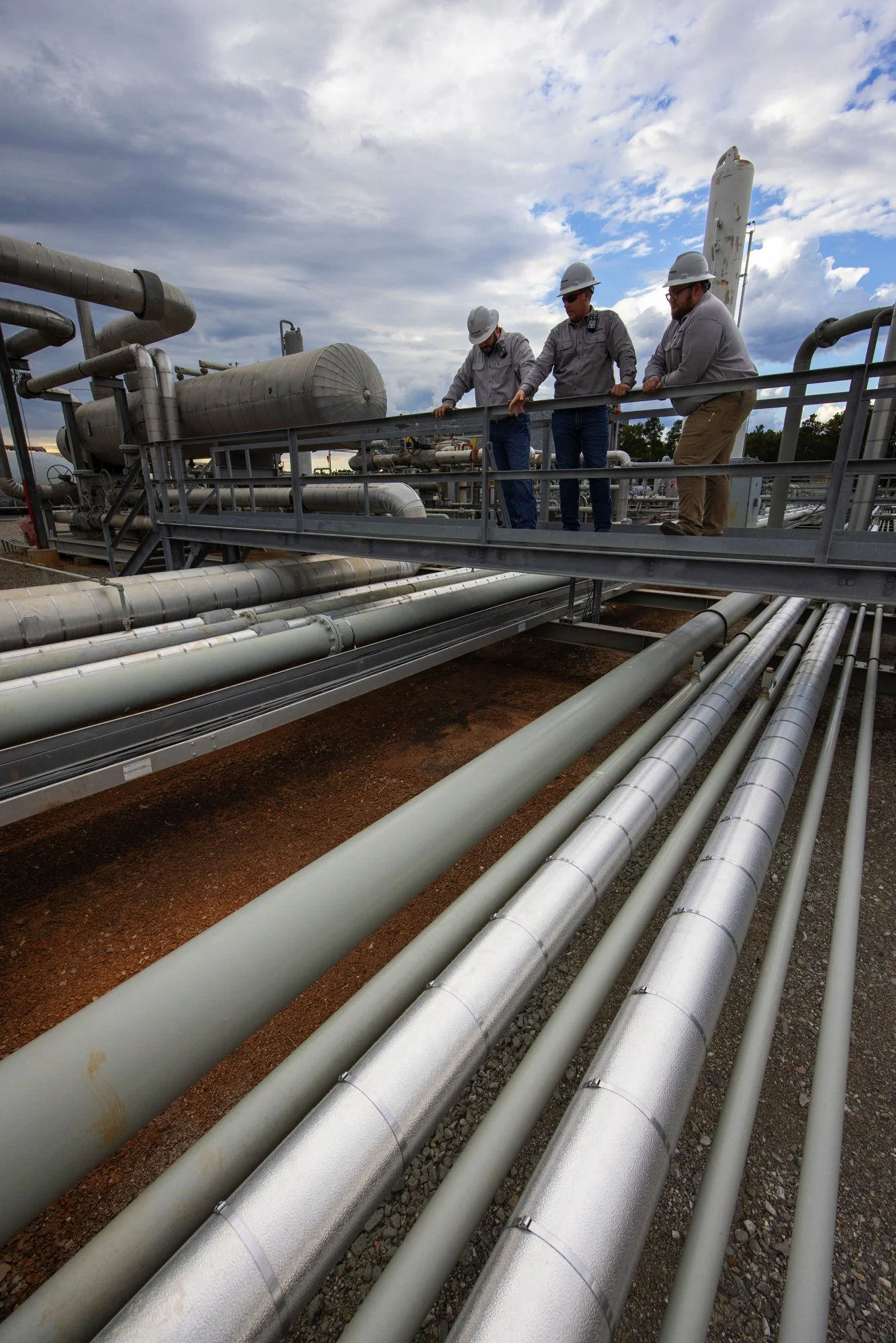 Three workers wearing hard hats, inspecting pipes and equipment on an industrial facility's outdoor platform with a cloudy sky in the background.