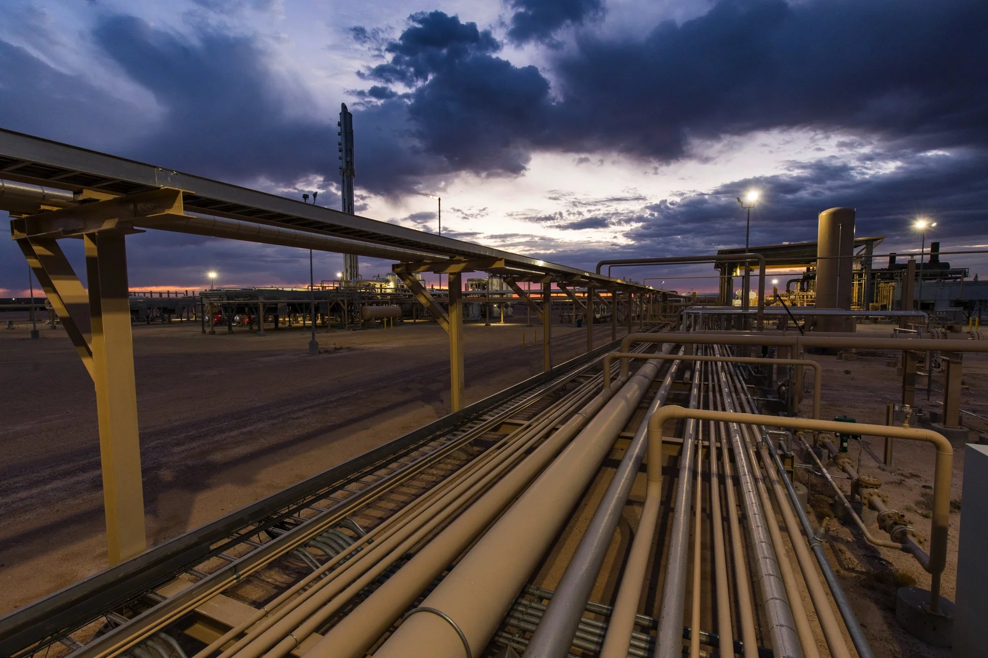 An industrial oilfield site with multiple pipelines and structures under a cloudy sunset sky.