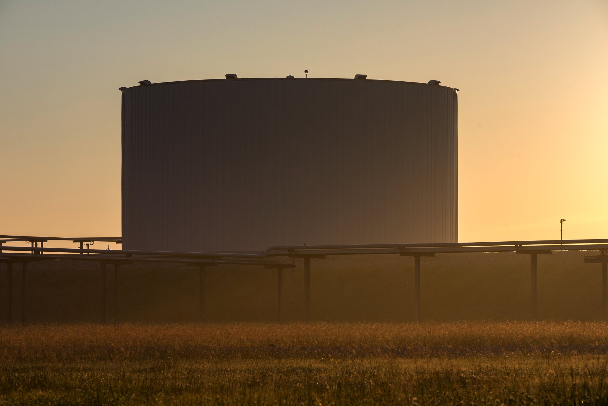 Large industrial storage tank or silo with a circular shape, surrounded by pipes and set against a sunset or sunrise sky.