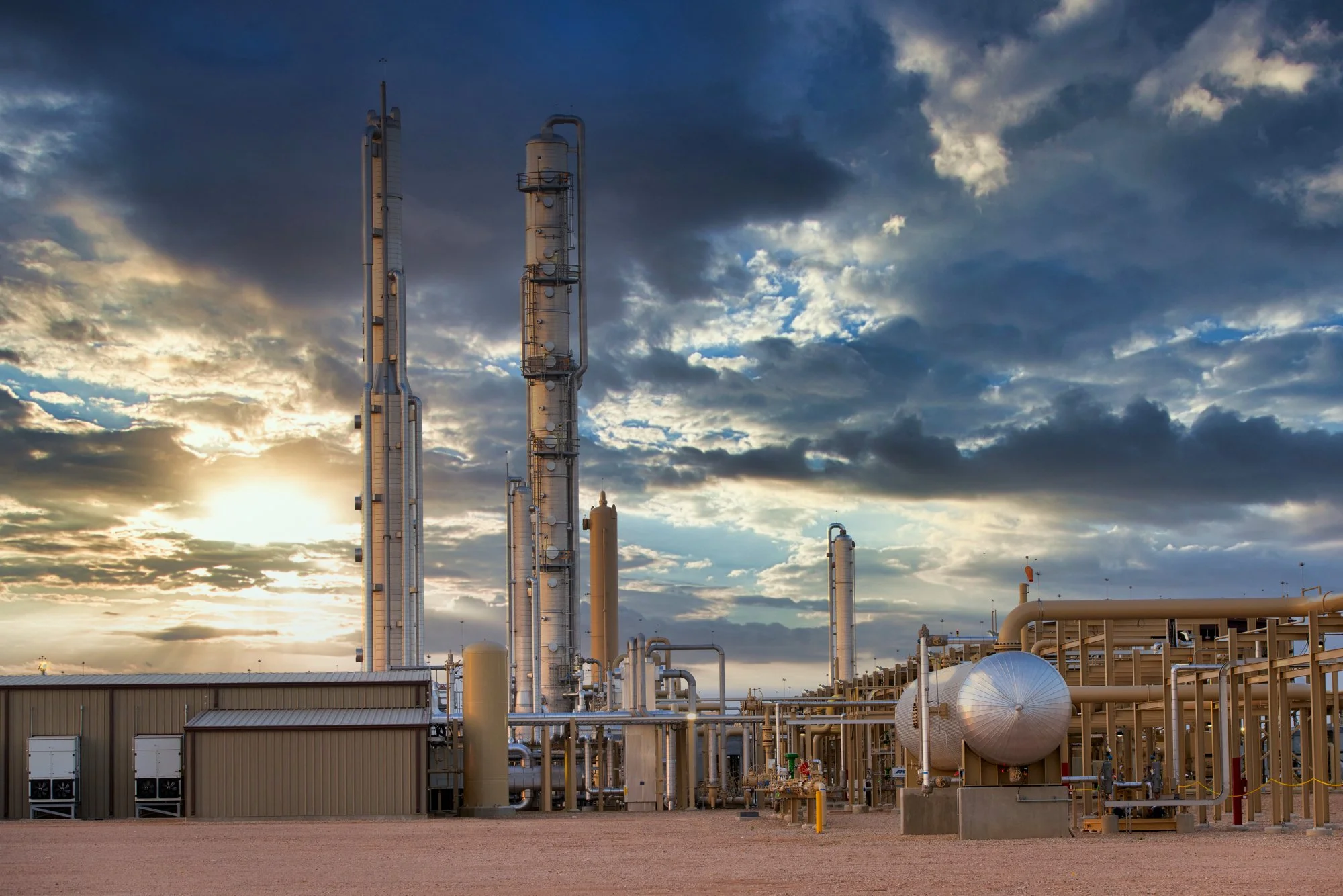 An industrial oil refinery with tall distillation towers and pipelines under a cloudy sky at sunset.