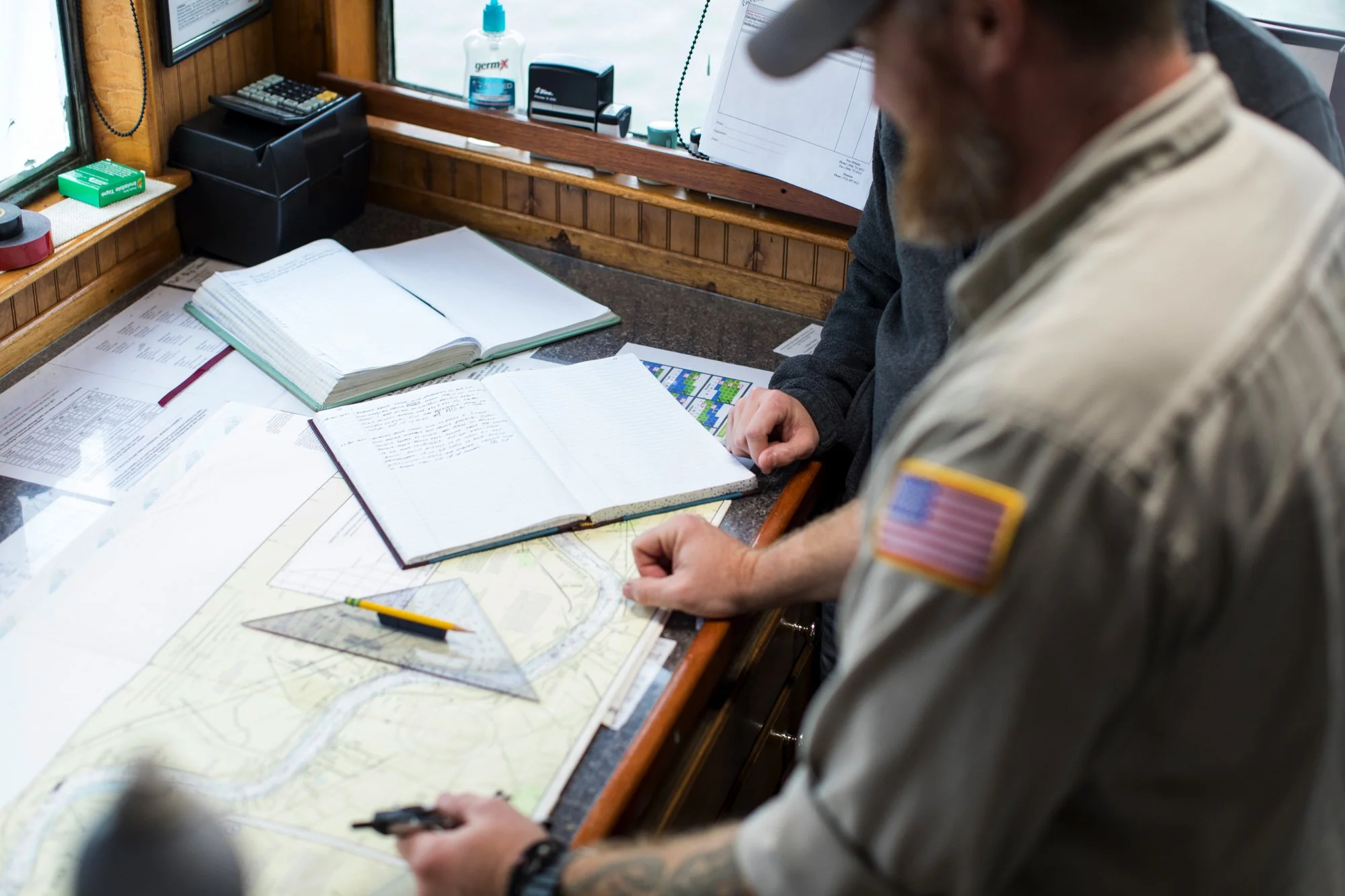 Two men studying maps and documents on a desk, with one wearing a uniform with an American flag patch on the sleeve.