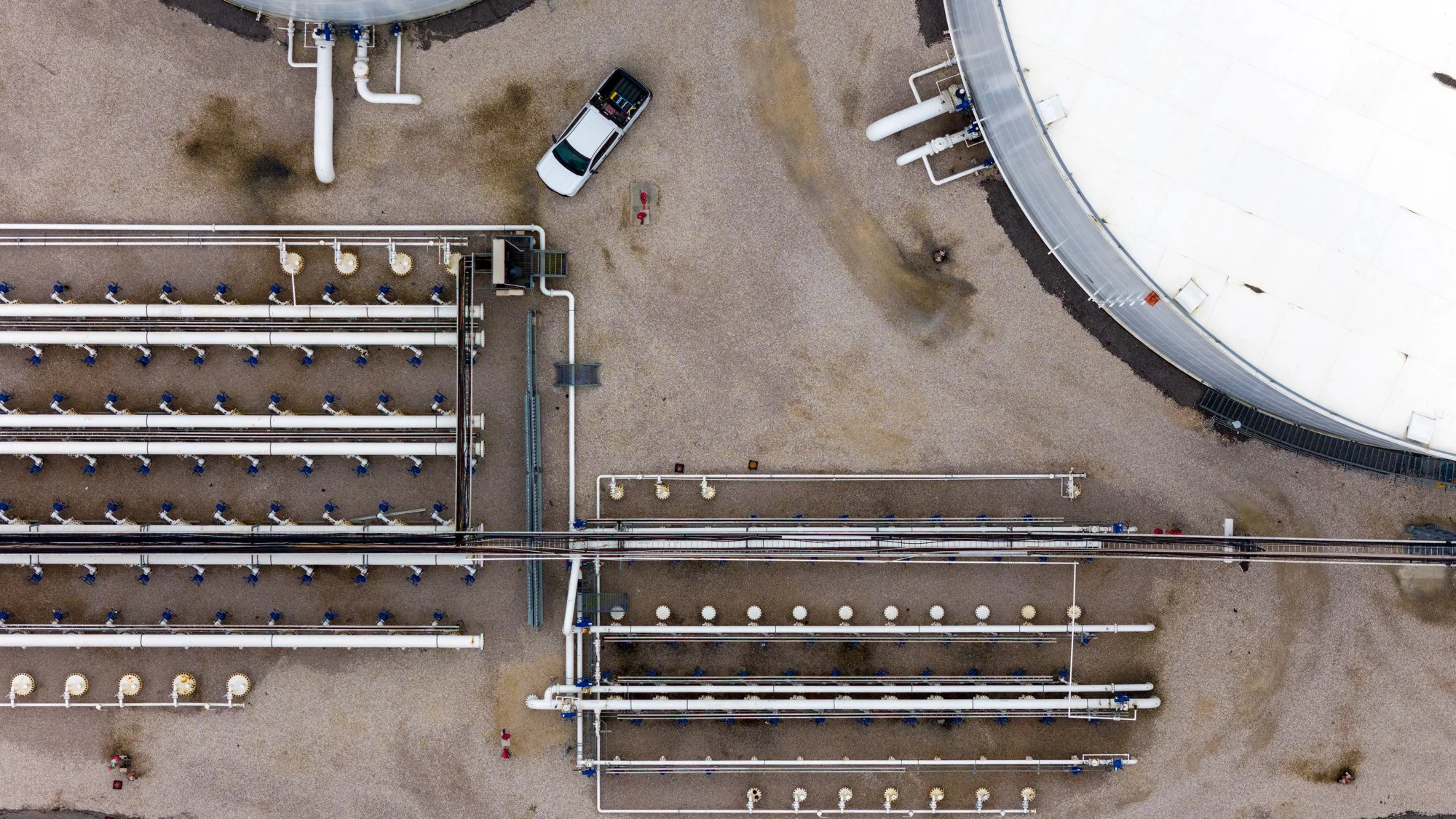 An aerial view of an industrial facility with large white tanks, pipelines, a white vehicle, and small red fire hydrants.