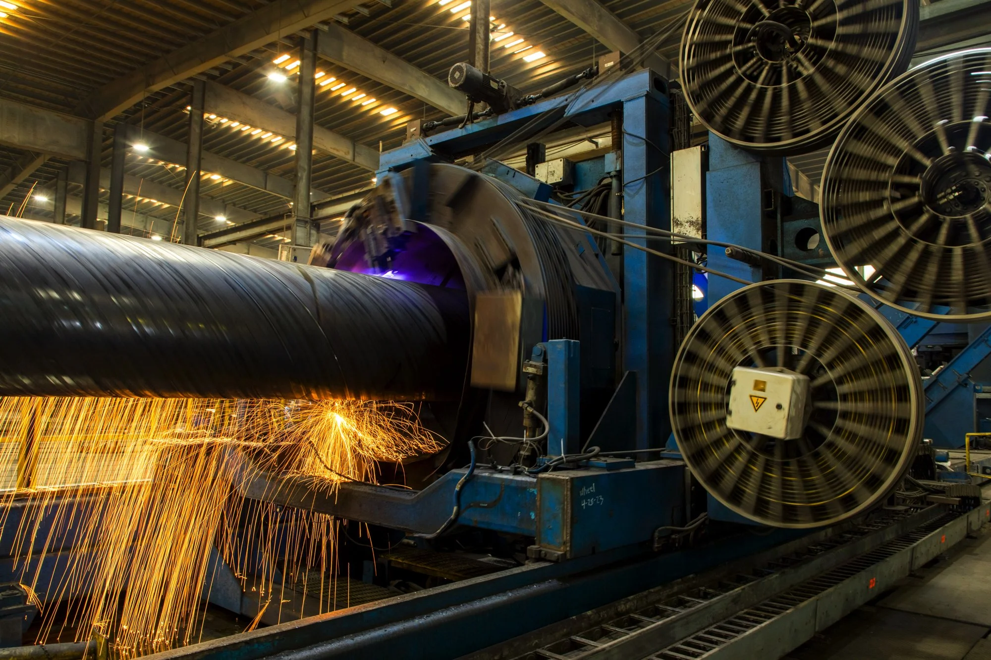Industrial manufacturing machine cutting or welding a thick steel pipe, with sparks flying, inside a factory.
