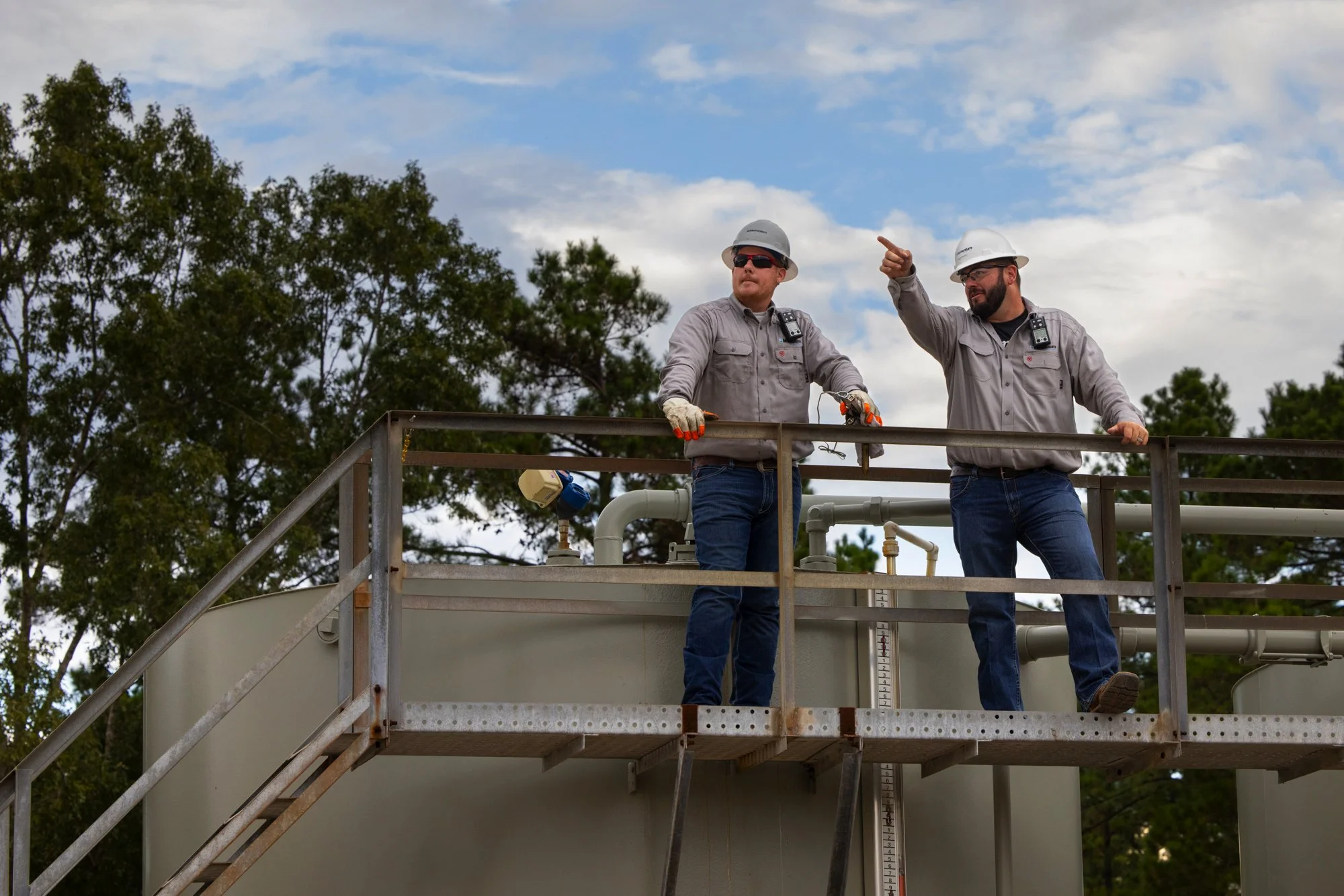Two workers wearing safety helmets and gray shirts standing on an industrial platform, discussing or inspecting equipment, with trees and a partly cloudy sky in the background.