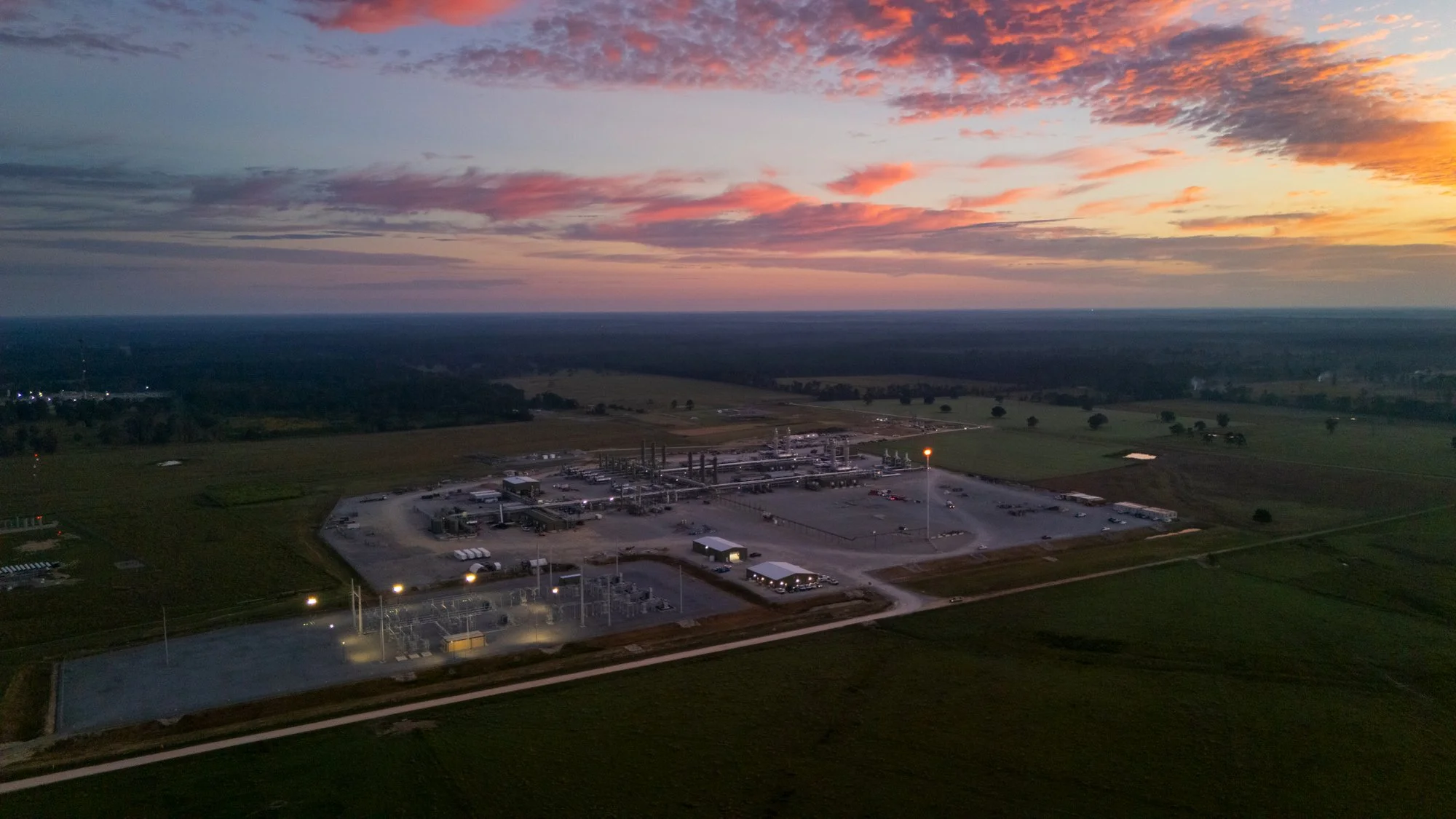 A gas station and industrial facility on a flat landscape during sunset with a colorful sky and scattered clouds.