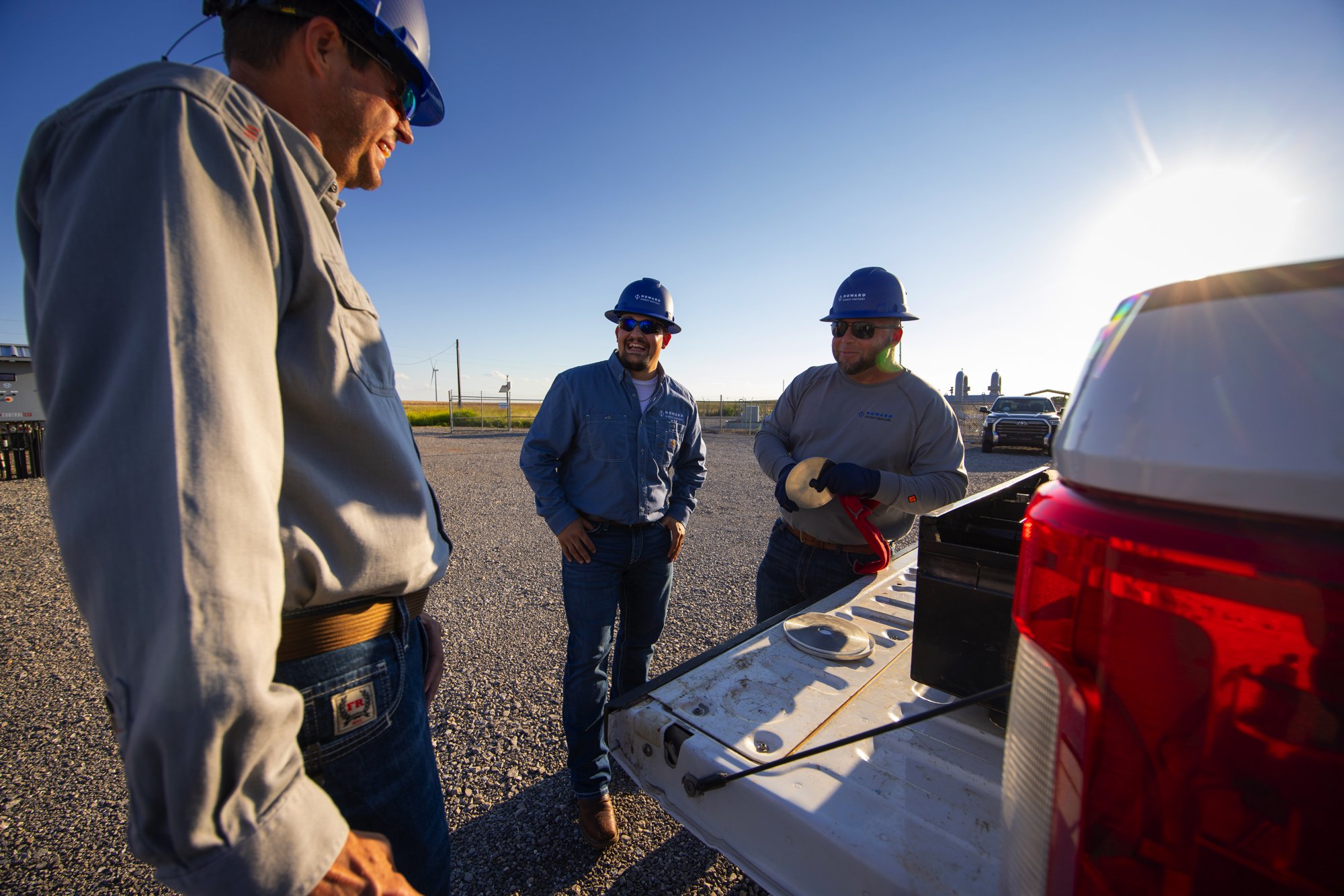 Three men wearing blue hard hats and safety glasses discussing near the back of a white truck in a gravel lot during sunset.