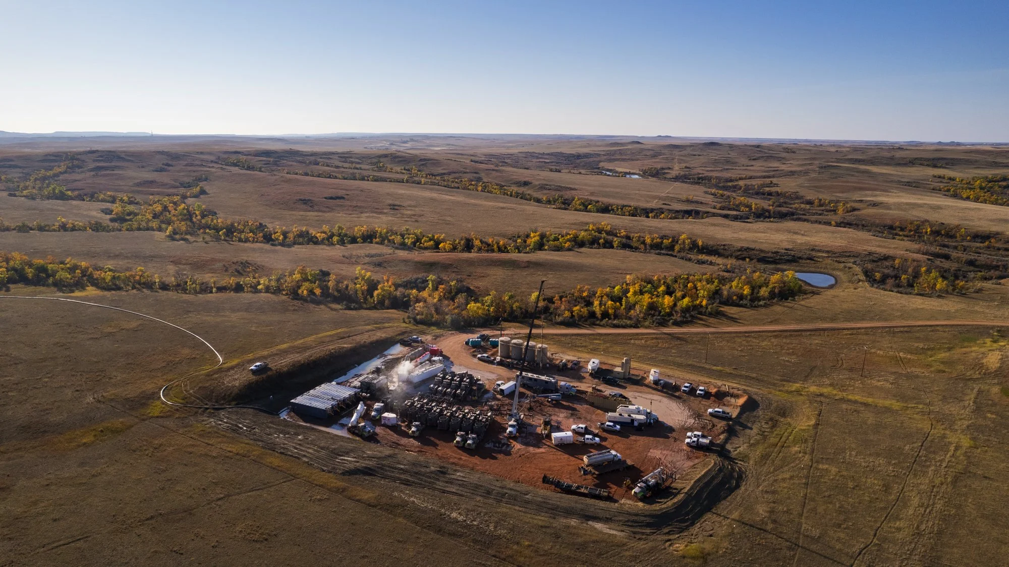 Aerial view of a large industrial drilling site in a rural landscape with fields and sparse trees, showing multiple trucks, machinery, and a tall drilling rig.