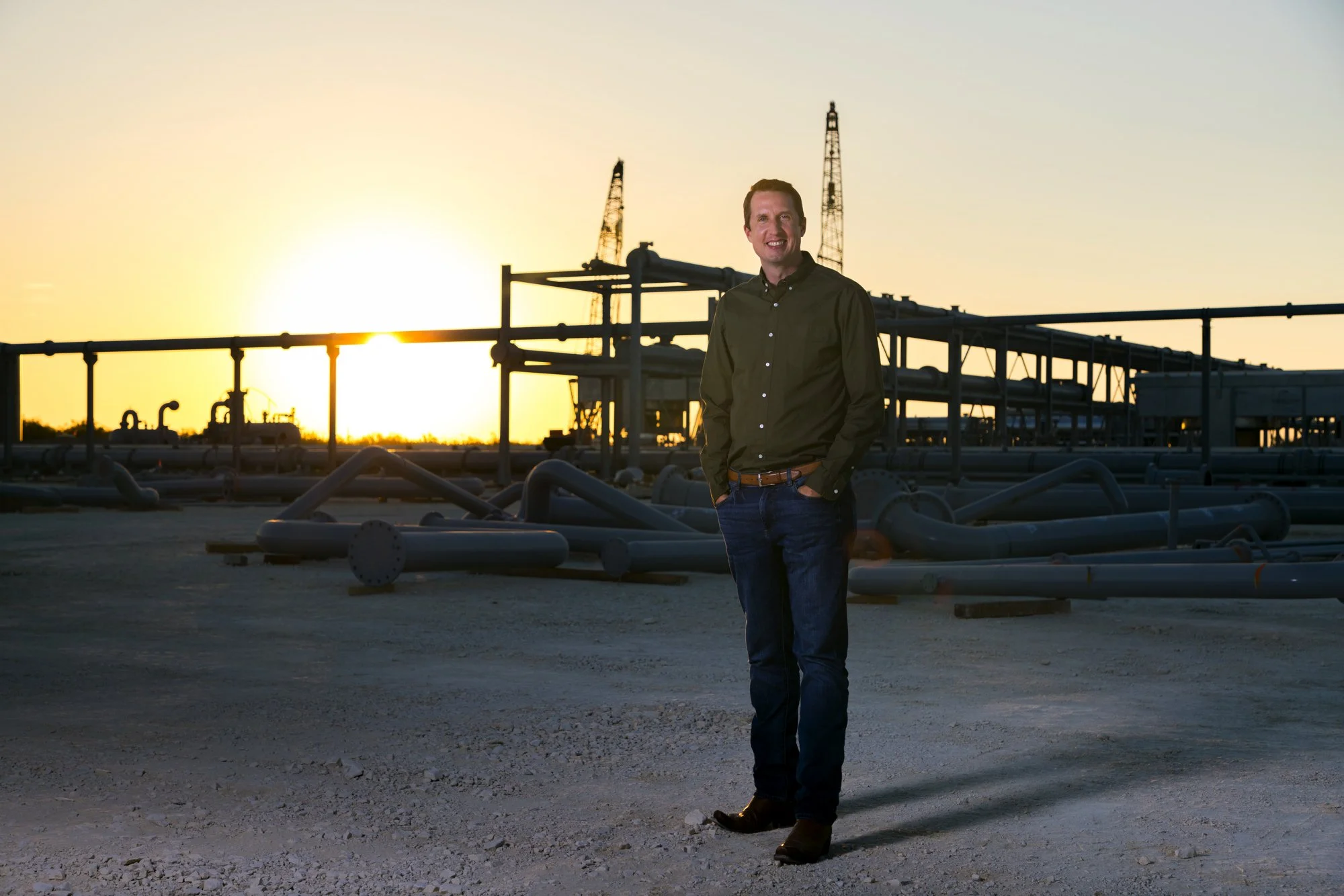A man standing outdoors at an oil or gas facility during sunset, surrounded by pipelines and industrial equipment.