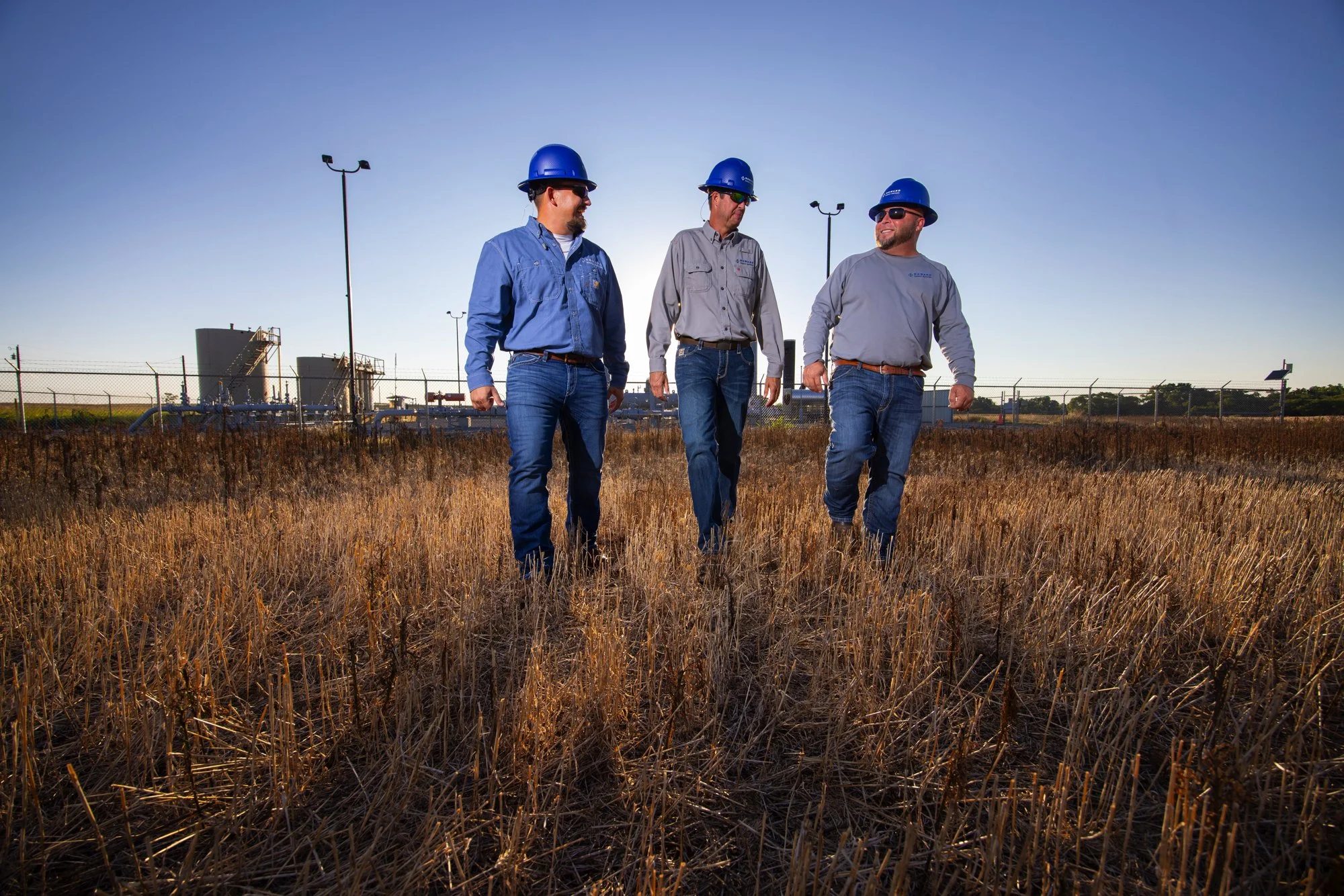 Three men wearing protective helmets and jeans walking through a dry field with industrial buildings in the background.
