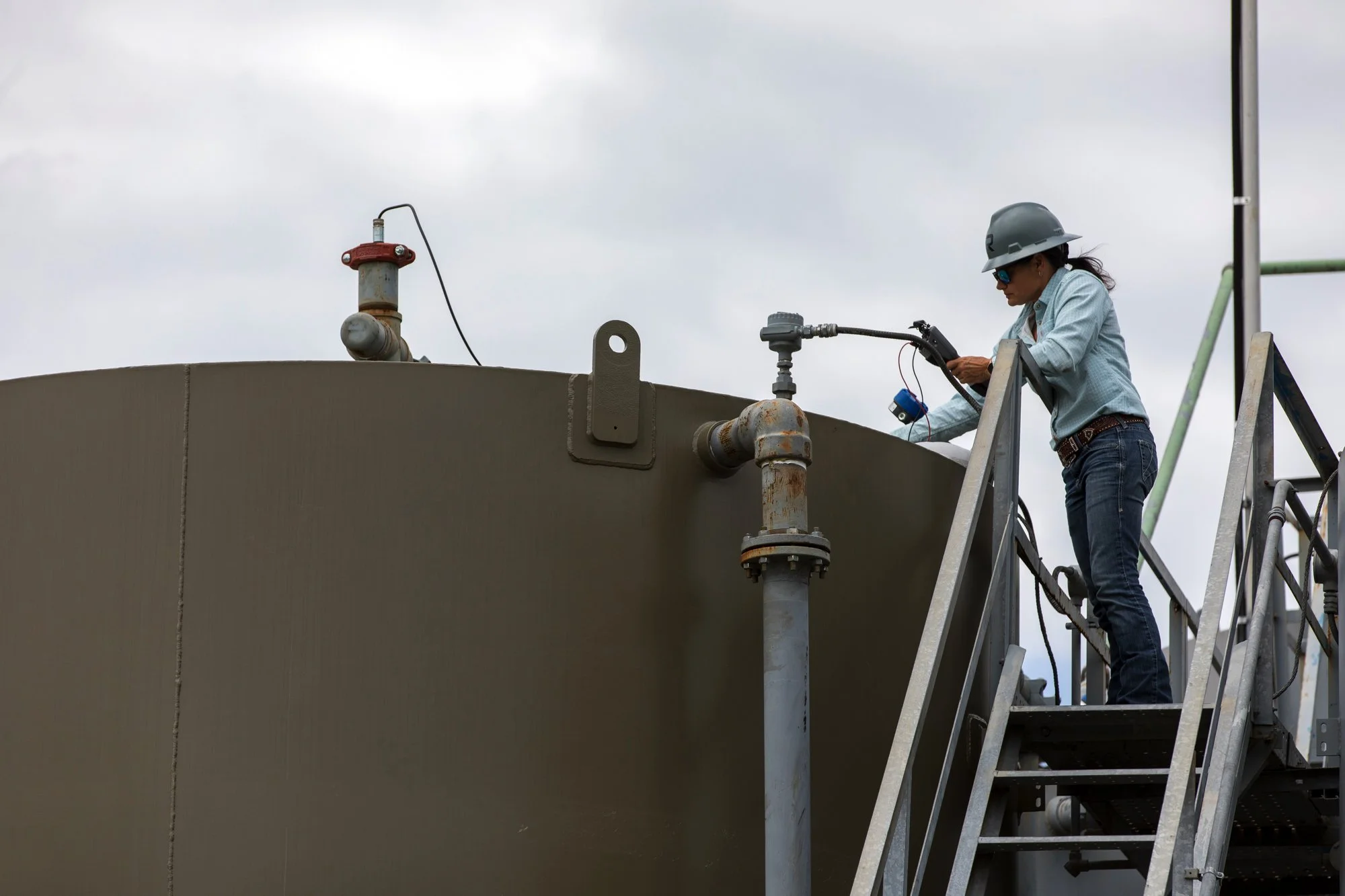 A woman wearing safety gear, including a gray hard hat and sunglasses, standing on a metal staircase examining or working with piping on a large industrial tank outdoors.