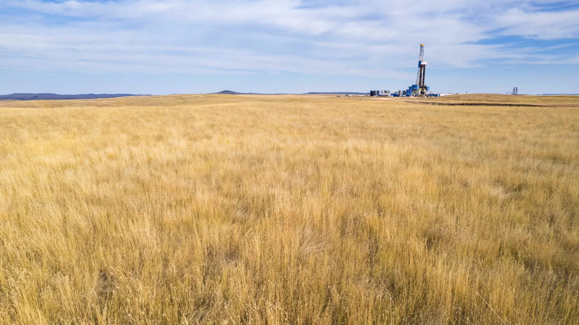 An expansive field of tall, golden grass with a distant oil rig drilling platform near the horizon under a partly cloudy sky.