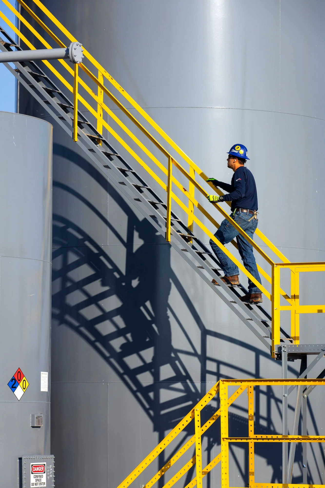 A worker in safety gear is ascending industrial stairs alongside a large gray tank, with shadows cast on the tank's surface.