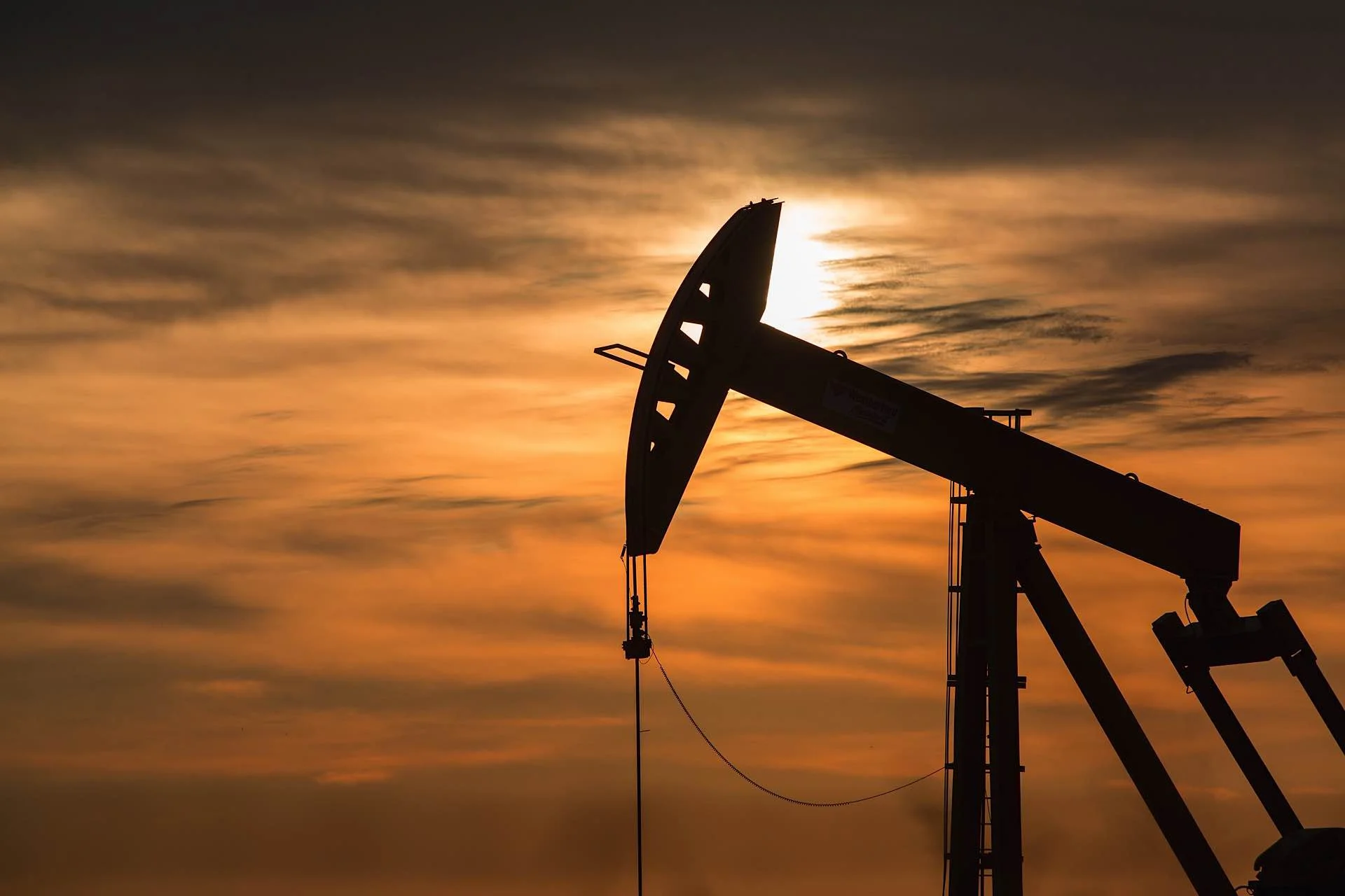 Silhouette of an oil pumpjack against a sunset sky with orange and yellow clouds.