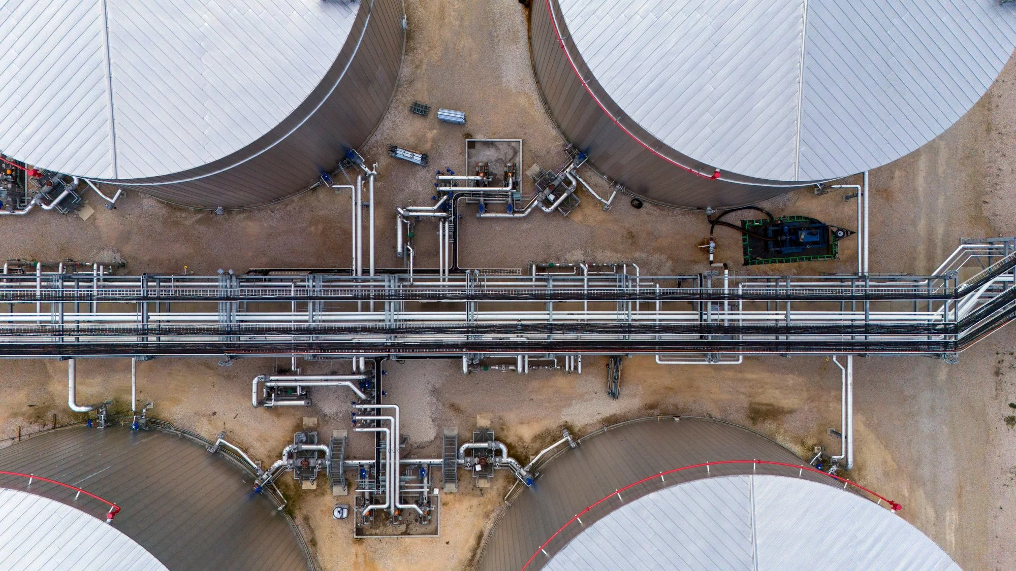 An aerial view of an industrial storage facility with large metal tanks, interconnected pipes, and a small forklift on the ground.